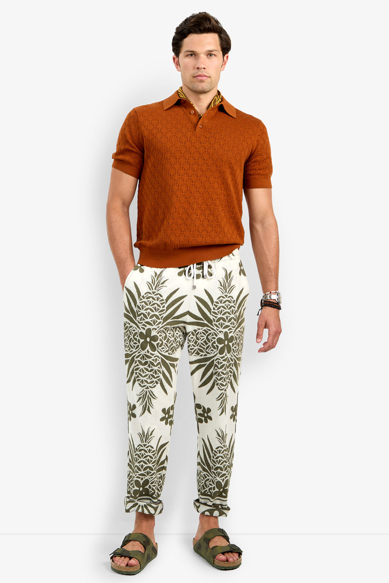 A man faces forward in a Waylen Short Sleeve Polo in teak, patterned drawstring pants with green and white tropical designs, green sandals, and bracelets on his left wrist. He has a neutral expression.