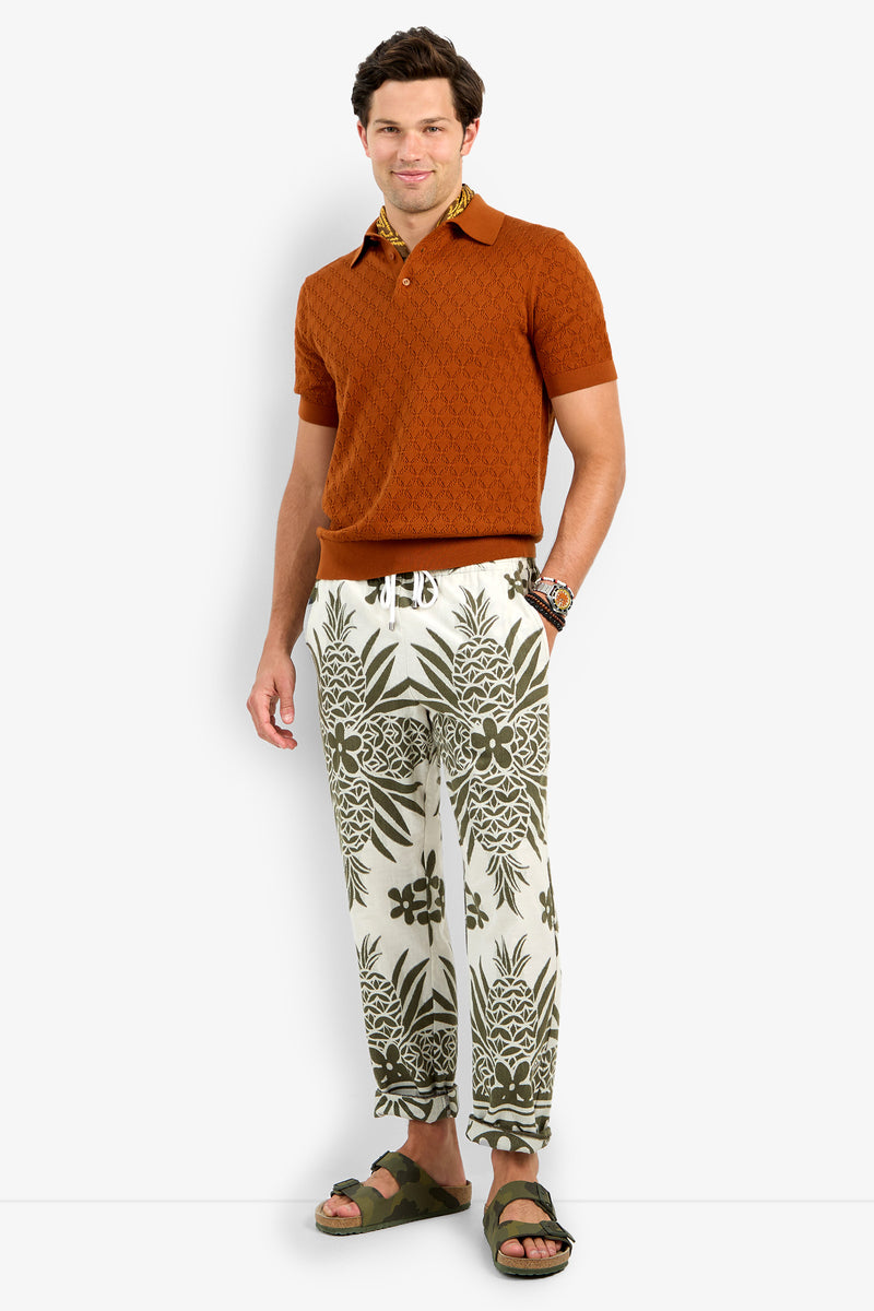 A man stands smiling, wearing a Waylen Short Sleeve Polo in brown, white and olive green drawstring pants with pineapples and flowers, olive sandals, and a brown patterned scarf around his neck.