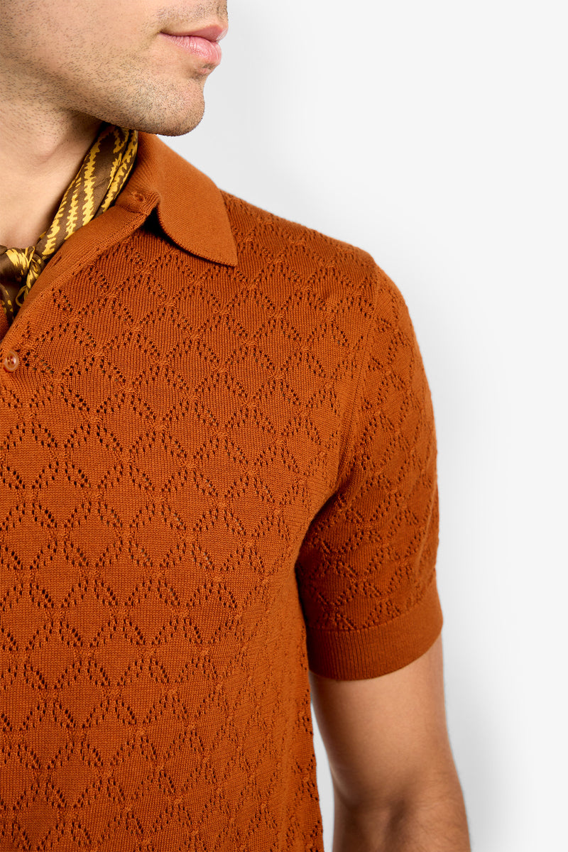 A man shown from the shoulders up against a plain background wears the Waylen Short Sleeve Polo in burnt orange, featuring a subtle textured pattern, paired with a brown patterned neck scarf.