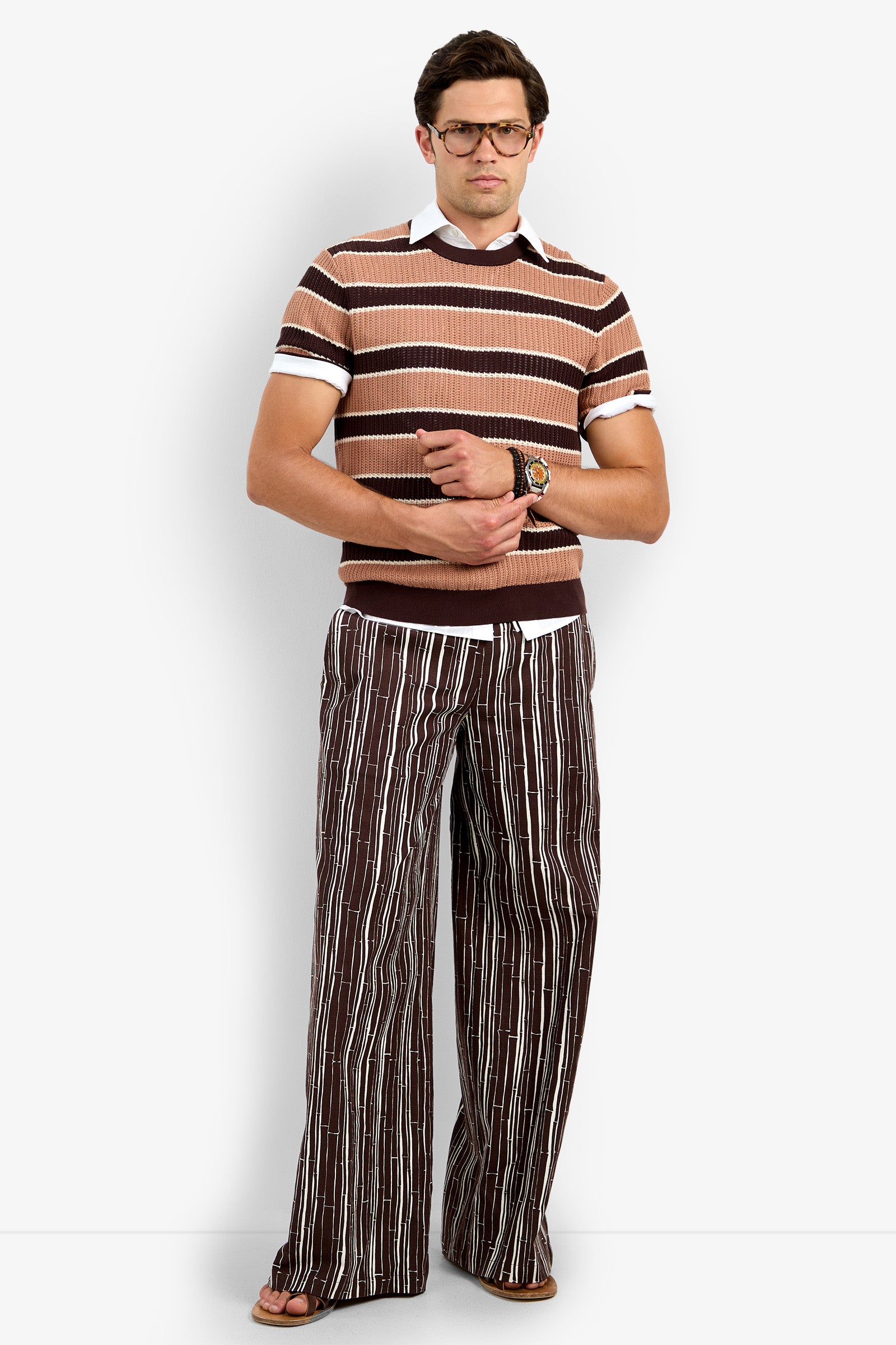 A man stands against a white background, wearing the Carlos Short Sleeve Crewneck, a brown and tan striped sweater over a white collared shirt, paired with brown striped wide-leg pants, brown sandals, glasses, and a wristwatch.