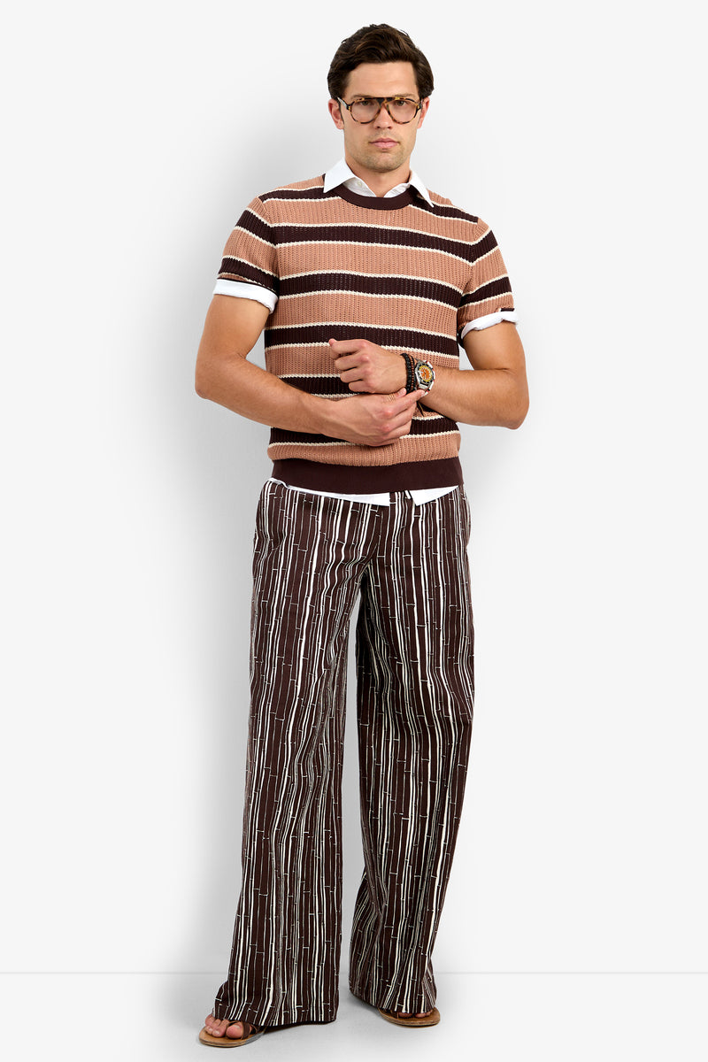 A man stands against a white background, wearing the Carlos Short Sleeve Crewneck, a brown and tan striped sweater over a white collared shirt, paired with brown striped wide-leg pants, brown sandals, glasses, and a wristwatch.
