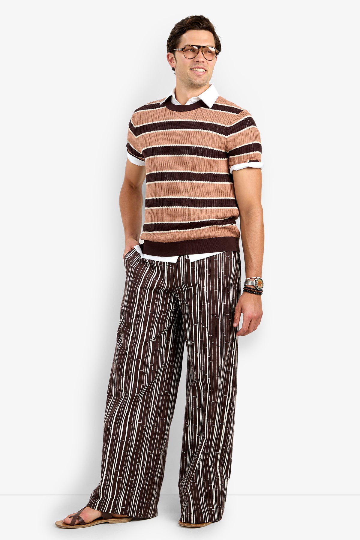 A man wearing glasses and the Carlos Short Sleeve Crewneck over a collared shirt, with wide-leg striped pants, sandals, and a bracelet, stands smiling with one hand in his pocket against a plain white background.