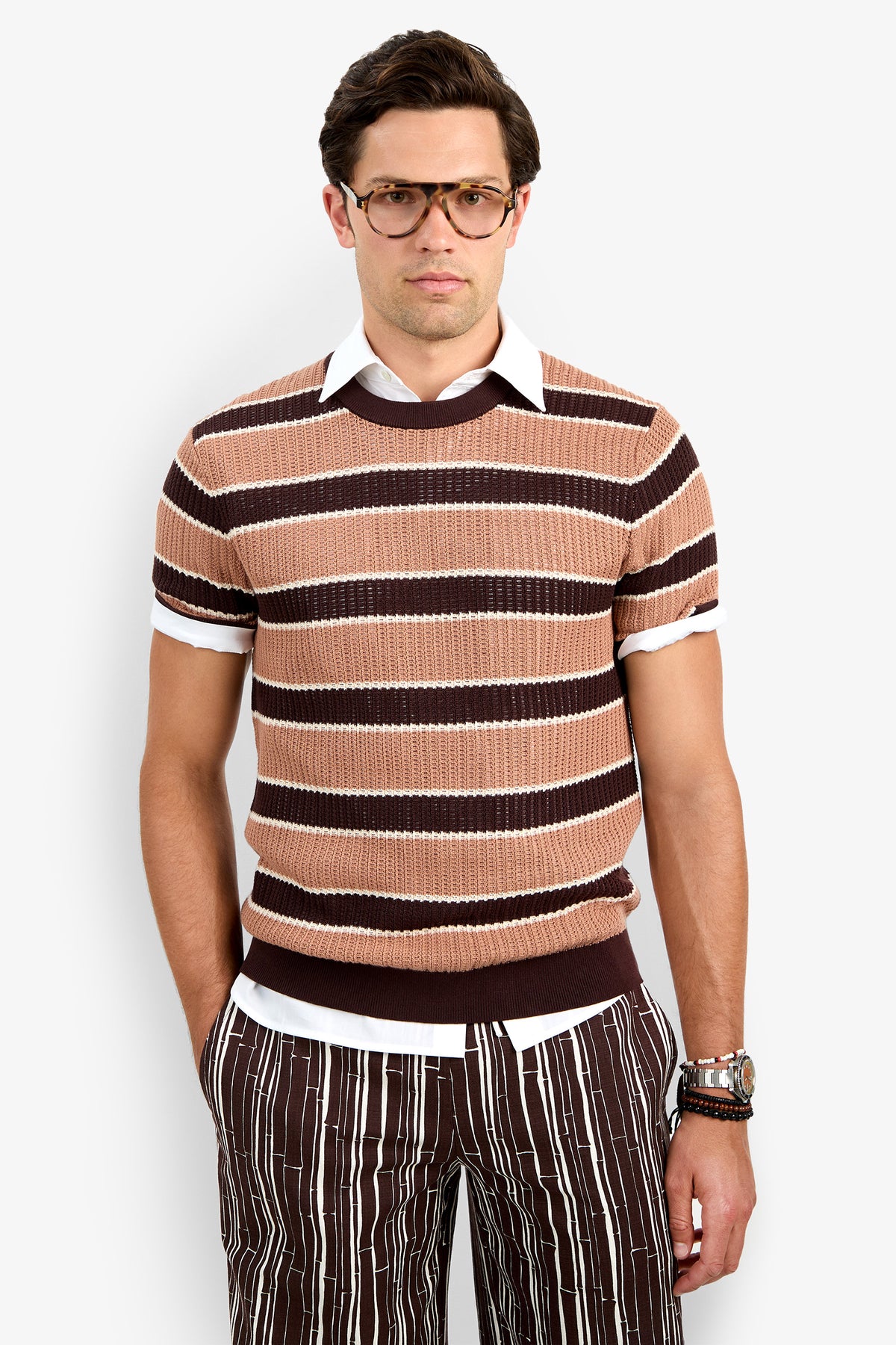 A man wears the Carlos Short Sleeve Crewneck over a white collared shirt and brown striped pants, standing with one hand in his pocket against a plain white background.