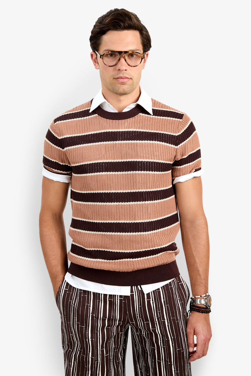 A man wears the Carlos Short Sleeve Crewneck over a white collared shirt and brown striped pants, standing with one hand in his pocket against a plain white background.