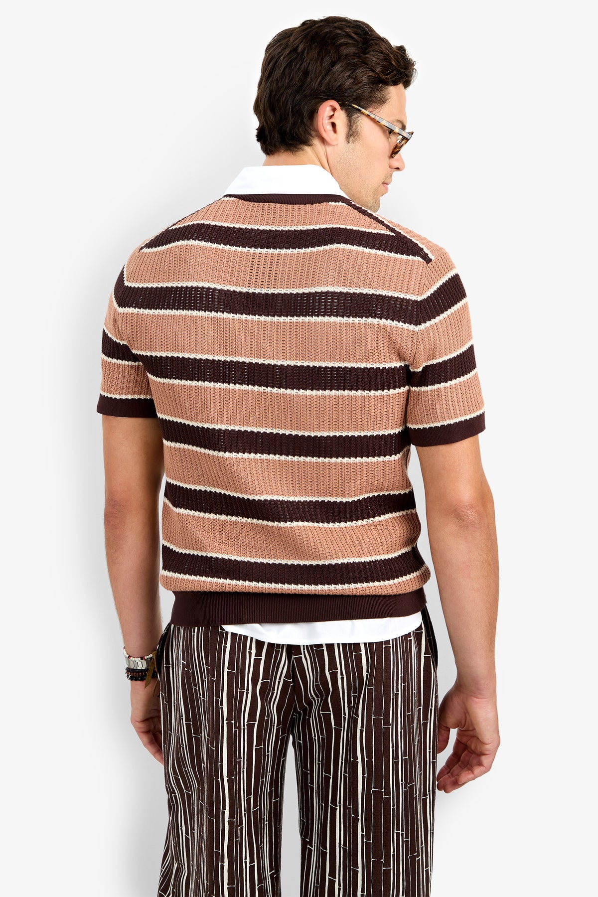 A man with short dark hair, glasses, and a wristwatch faces away, wearing the Carlos Short Sleeve Crewneck—a brown, beige, and white striped knit shirt—and brown pants with vertical white stripes.