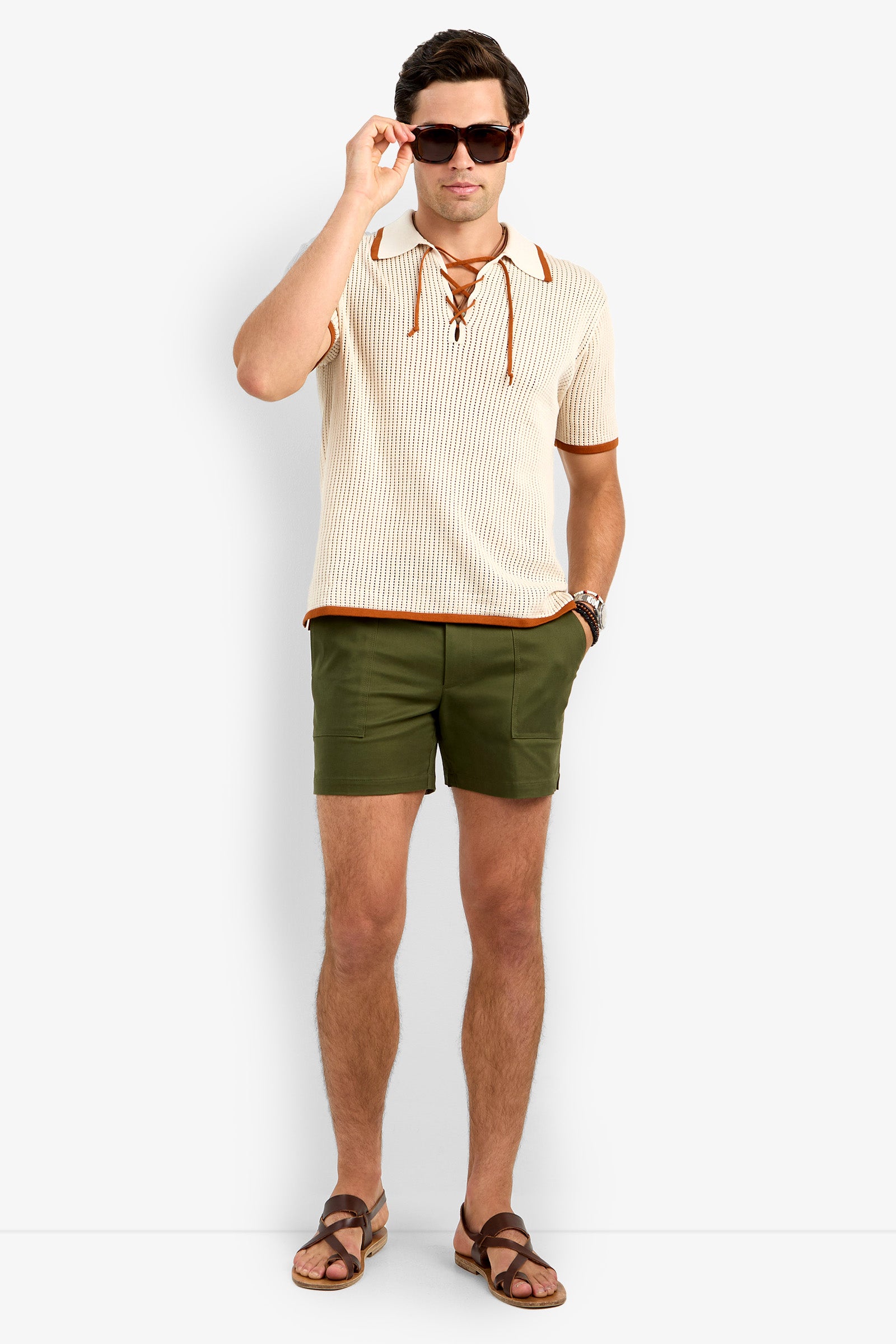 A man in sunglasses, the cream Simon Lace Up Polo with brown trim, olive green shorts, and brown sandals stands against a plain white background.