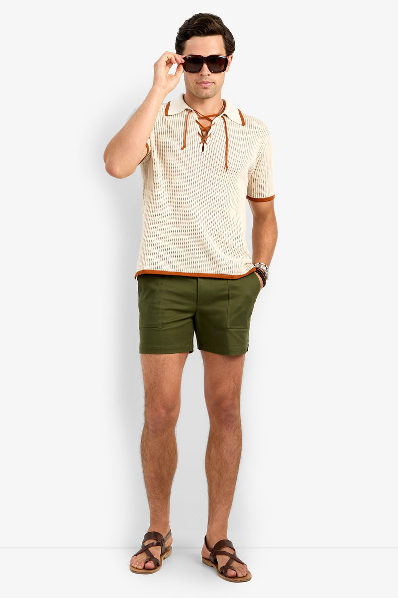 A man in sunglasses, the cream Simon Lace Up Polo with brown trim, olive green shorts, and brown sandals stands against a plain white background.