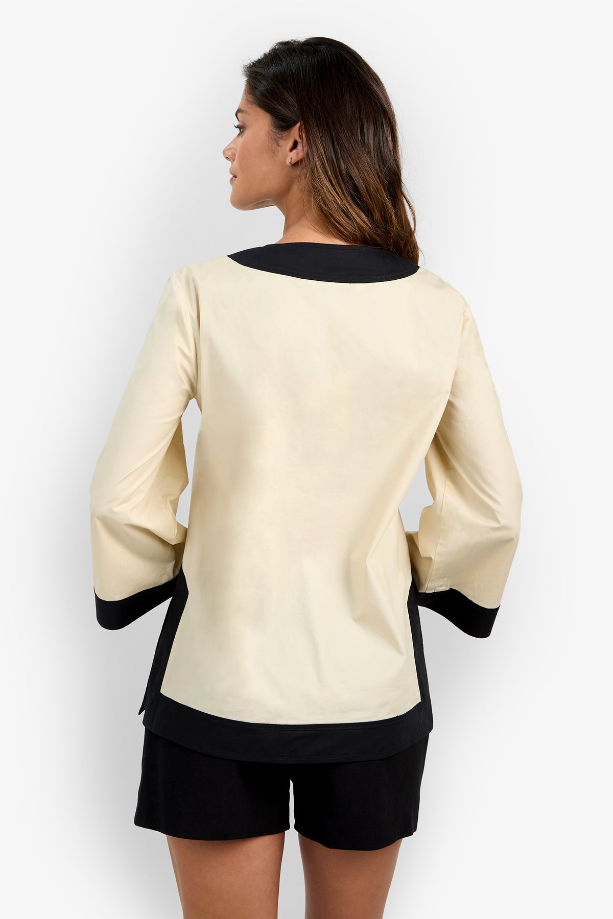 A woman is standing with her back to the camera, wearing the Dametria Top—a cream-colored long-sleeve top with black cuffs and collar—paired with black shorts.