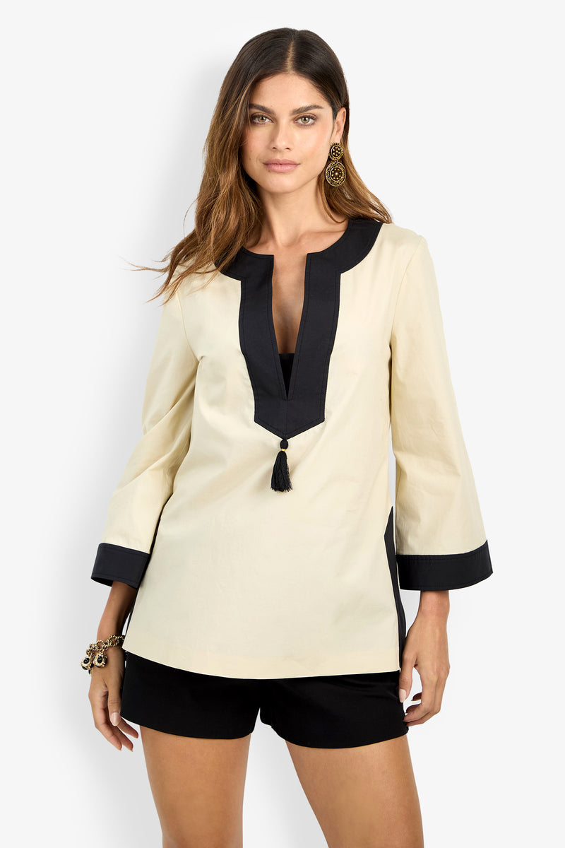 A woman with long brown hair models the DAMETRIA TOP, a cream tunic featuring black trim and a front tassel detail, paired with black shorts. She accessorizes with gold earrings and a bracelet, standing against a plain white backdrop.
