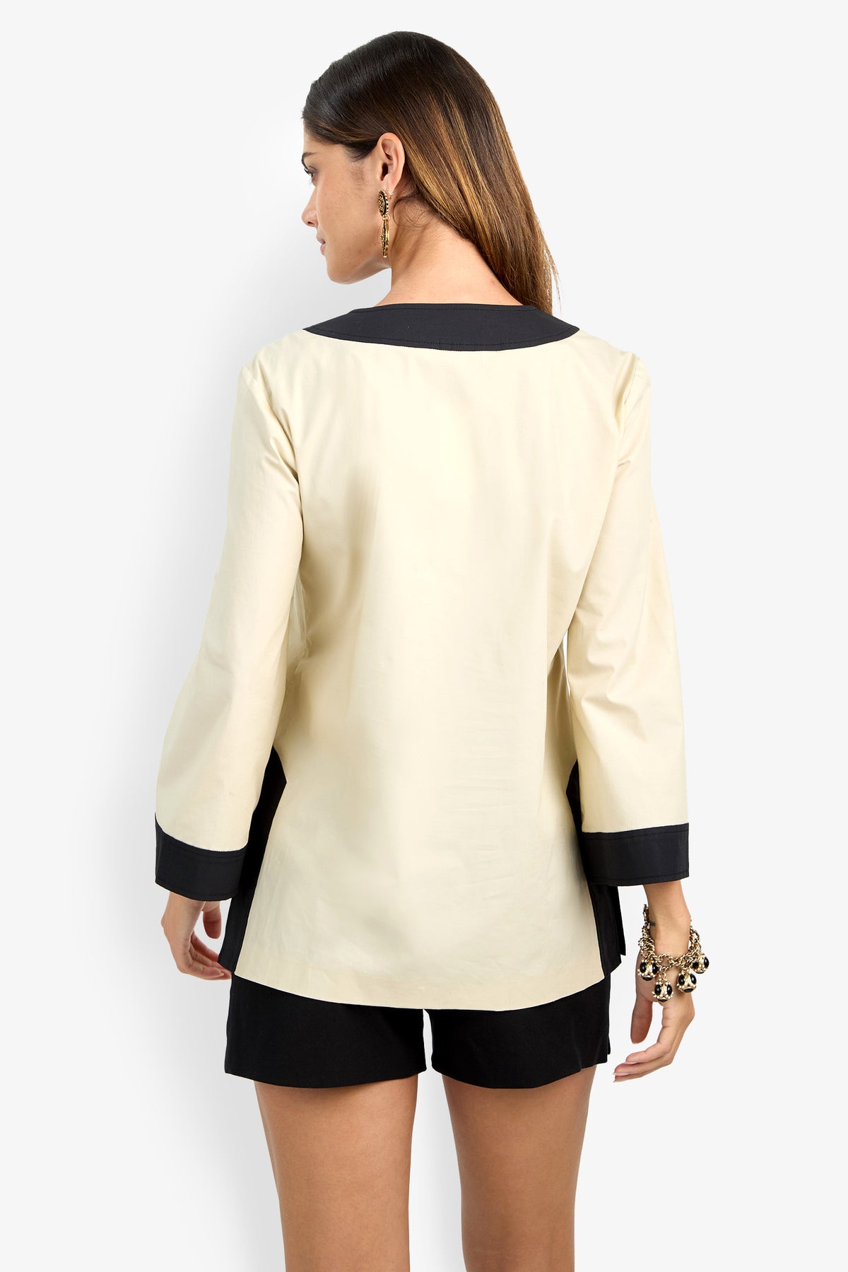 A woman with long brown hair, gold earrings, and black shorts models the DAMETRIA TOP—cream with black collar and cuffs—standing against a plain white background, facing away from the camera.