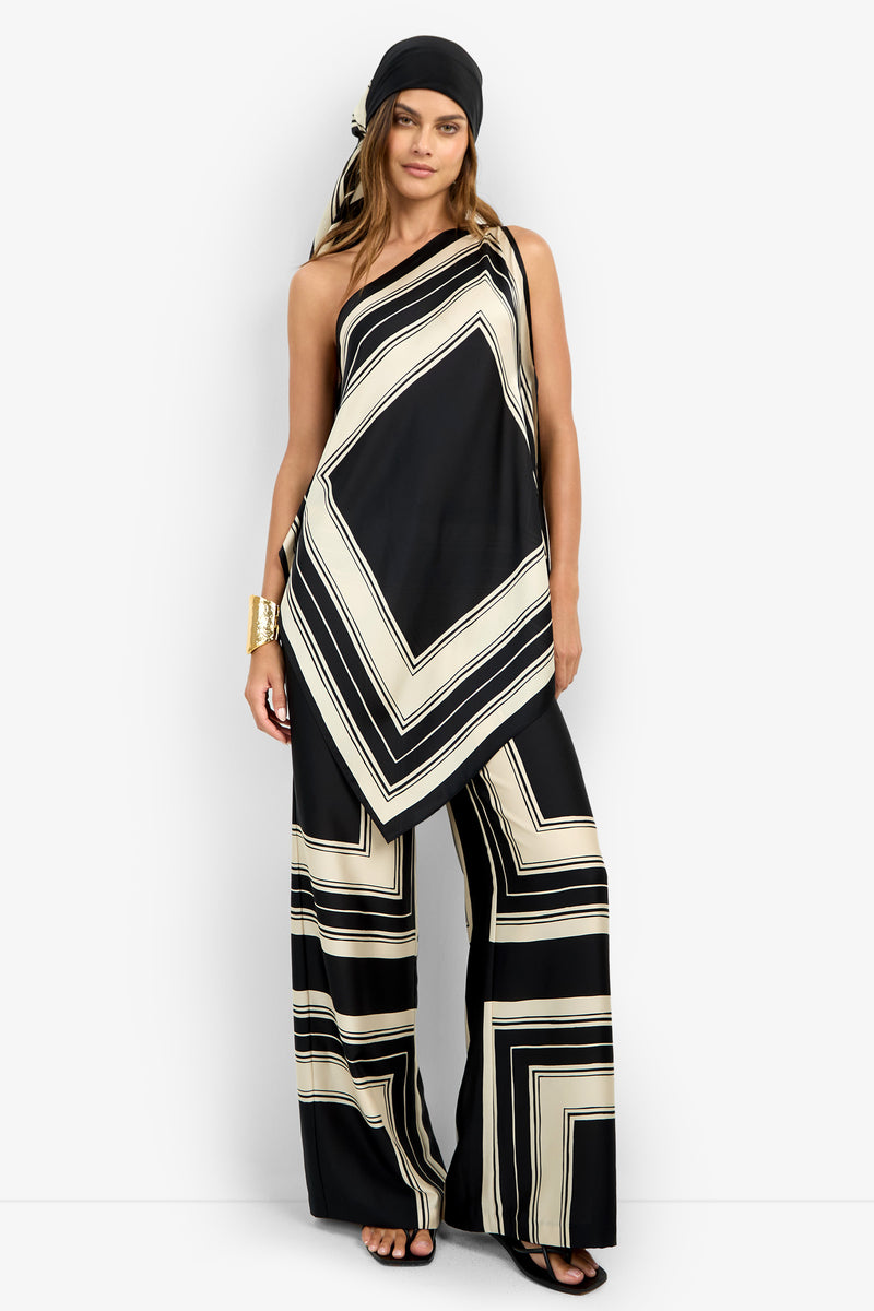 A woman wears the DIANN TOP—an asymmetrical black top with bold cream geometric stripes—paired with matching wide-leg pants, a black headscarf, black sandals, and a gold cuff bracelet.