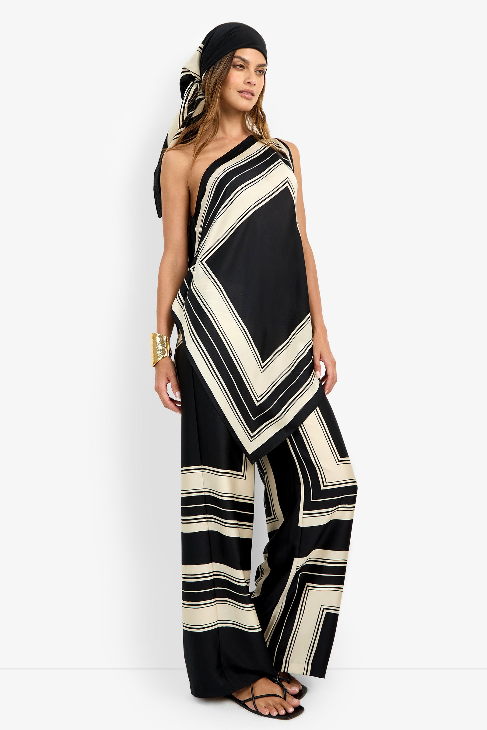 A woman wears the DIANN TOP—a black and cream geometric-patterned one-shoulder top—with matching wide-leg pants, paired with a black headscarf, gold cuff bracelet, and black sandals.