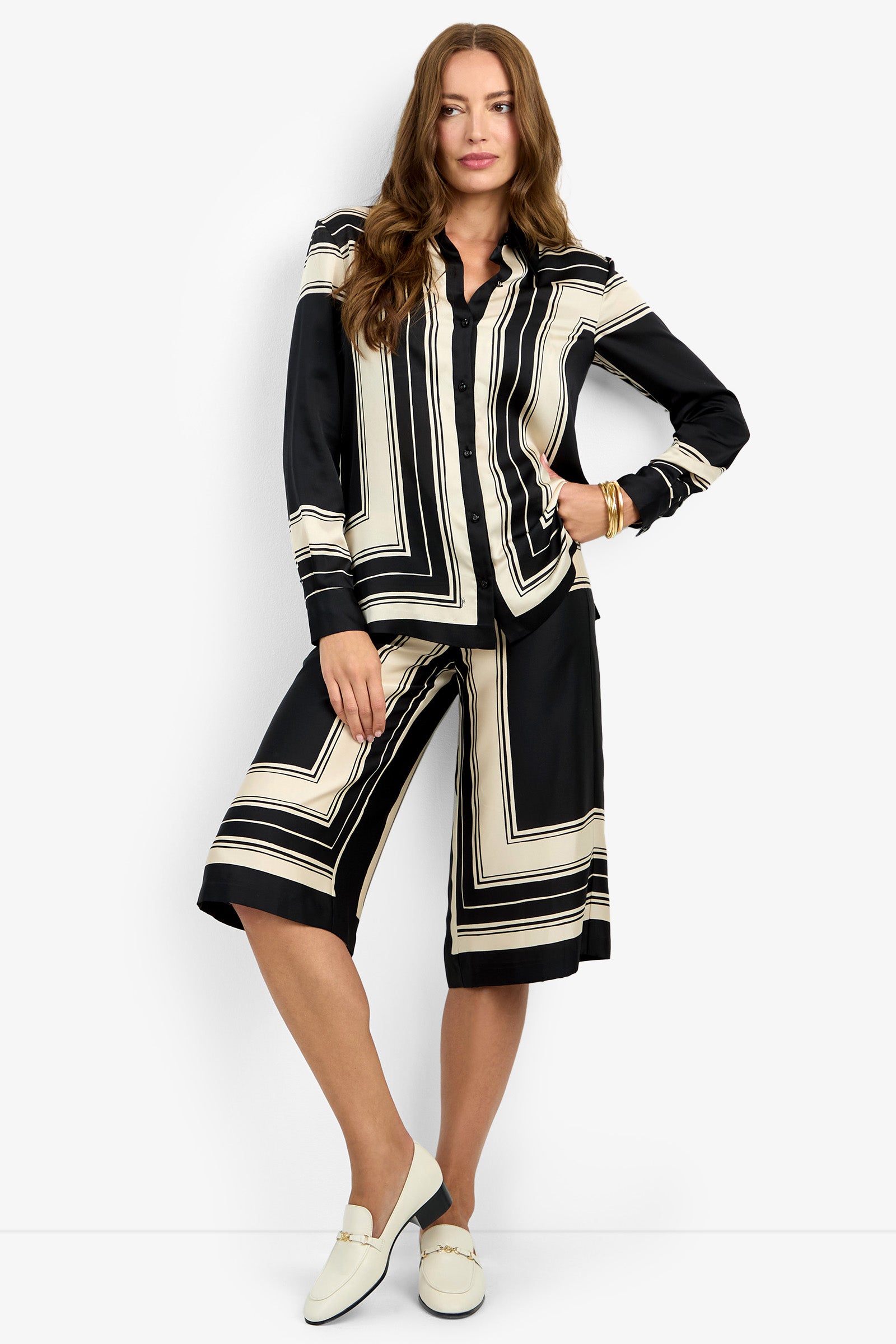 A woman with long, wavy hair poses confidently against a white background wearing the BRISTLECONE TOP—a black and cream geometric-patterned blouse—with matching knee-length shorts and cream loafers.