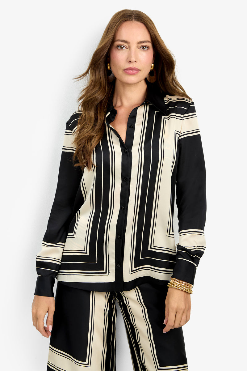 A woman with long brown hair wears the BRISTLECONE TOP, a black and cream geometric patterned button-up shirt, paired with matching pants. She accessorizes with gold hoop earrings and several gold bangles, standing against a plain white background.