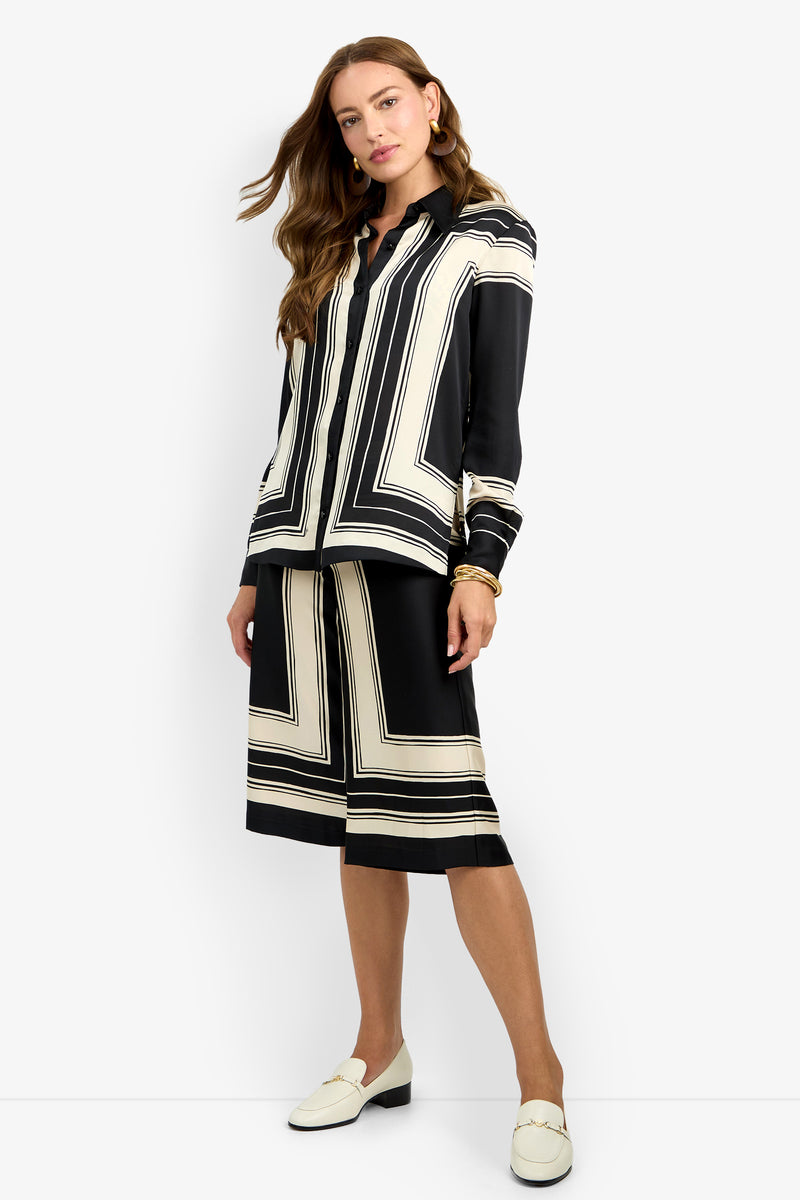 A woman with long wavy hair models the BRISTLECONE TOP, a black and white geometric patterned blouse, paired with a matching knee-length skirt and white loafers against a plain white background.