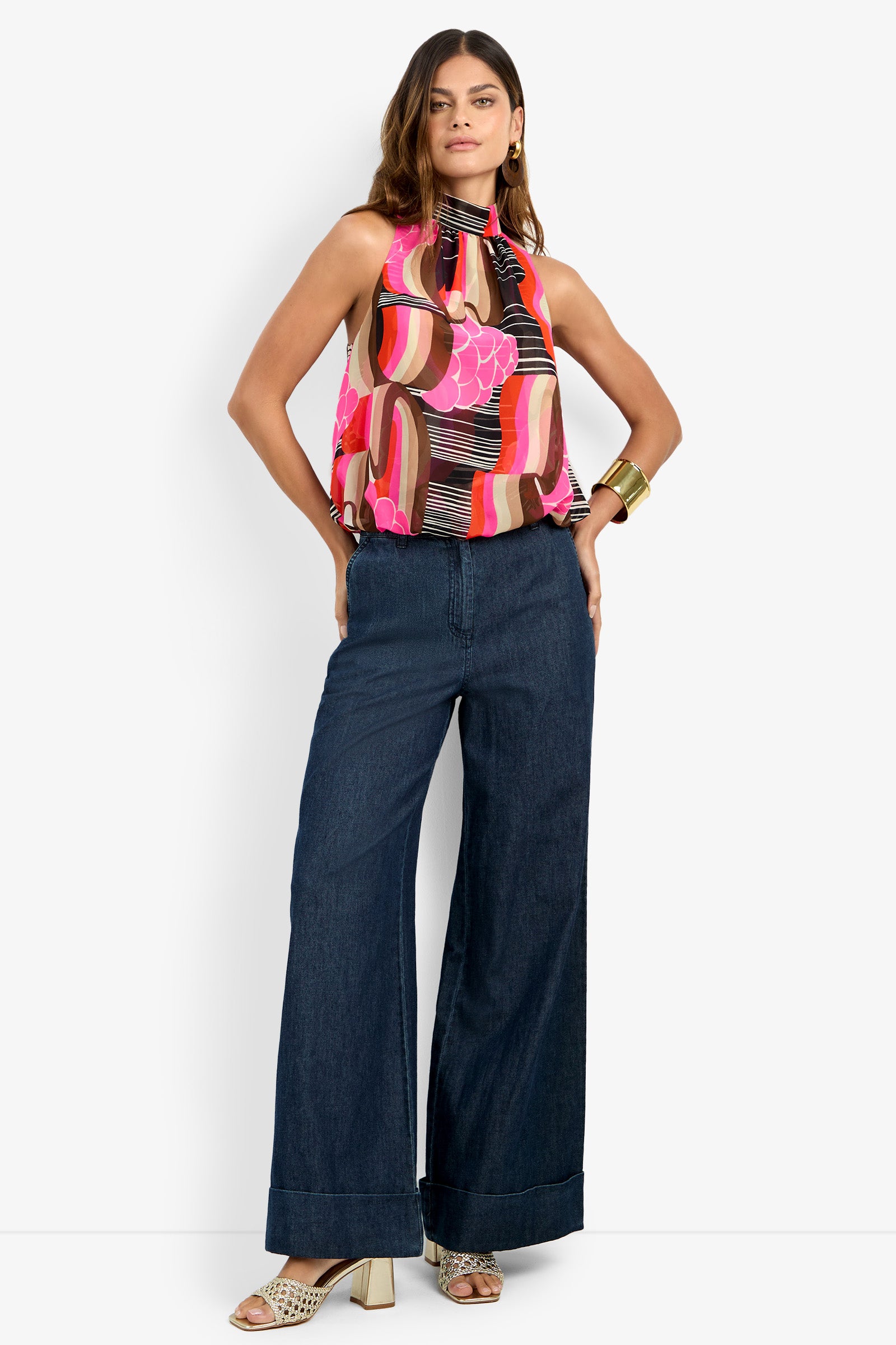 A woman stands confidently wearing the wide-leg DILLAN PANT in dark denim, paired with a colorful sleeveless halter top, heeled sandals, hoop earrings, and a large gold cuff bracelet.