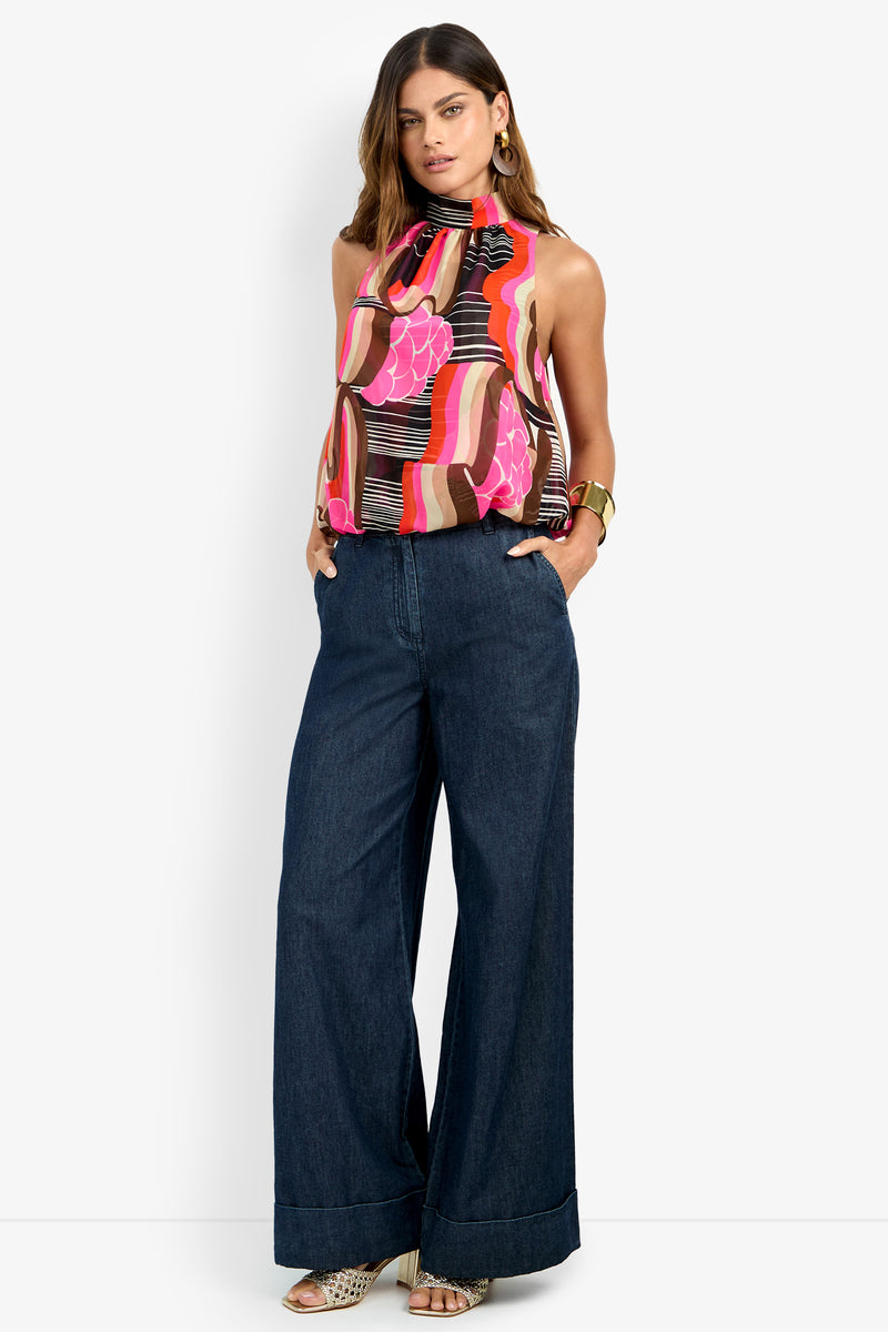 A woman stands with hands in her pockets, wearing the CORTONA TOP—a colorful sleeveless halter with pink, brown, and orange patterns—paired with wide-leg dark blue jeans, gold earrings, and beige heeled sandals.