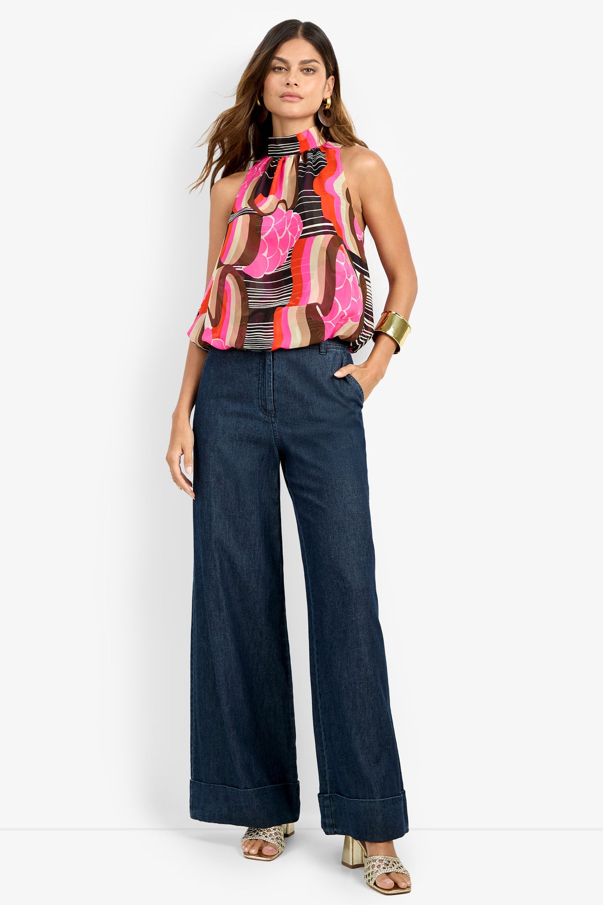 A woman stands with one hand in her pocket, wearing the colorful patterned CORTONA TOP, wide-leg dark blue jeans, gold hoop earrings, a chunky bracelet, and heeled sandals.