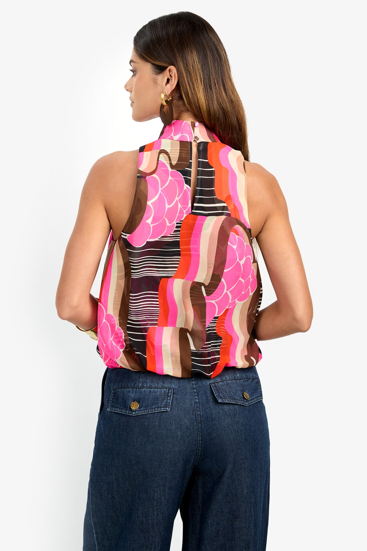 A woman with straight brown hair and gold hoop earrings faces away, wearing the sleeveless CORTONA TOP in a bold pink, red, brown, and white abstract print, paired with high-waisted blue jeans.
