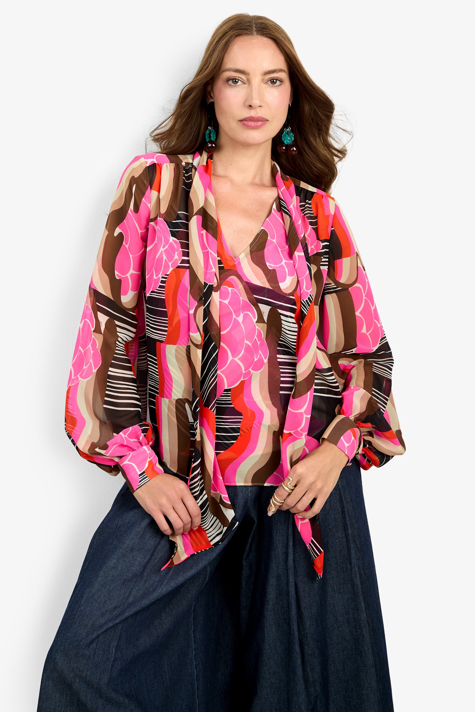 A woman with long brown hair wears the PIENZA TOP, a colorful patterned blouse featuring pink, brown, orange, and black designs. She pairs it with wide-leg dark denim pants and poses with one hand in her pocket.