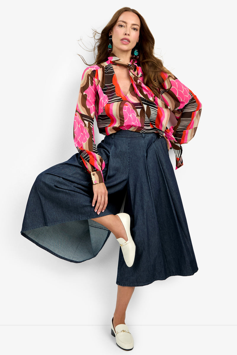 A woman poses with one leg raised, wearing the PIENZA TOP—a colorful, patterned blouse in pink, brown, and orange—paired with a denim midi skirt, white loafers, and statement earrings against a plain background.