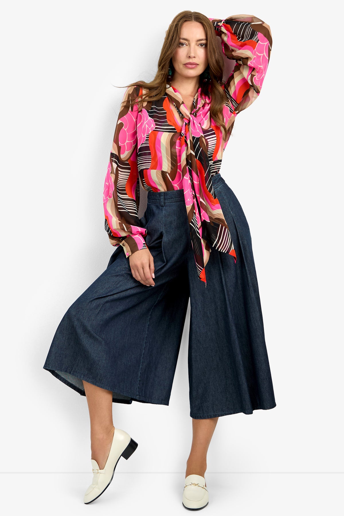 Woman wearing the PIENZA TOP—a colorful, patterned long-sleeve blouse with a bow tie neck—paired with wide-leg dark denim culottes and white loafers, standing against a plain white background.
