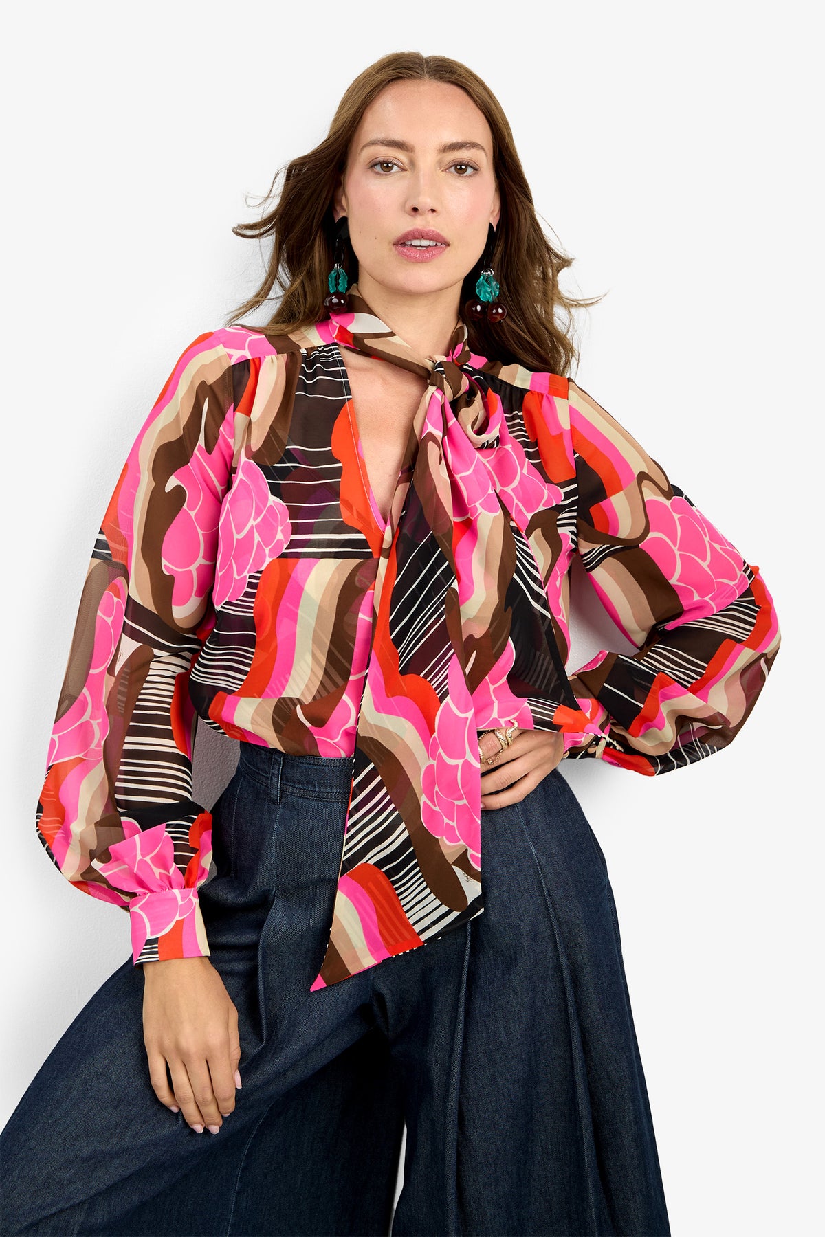 A woman wears the PIENZA TOP—a colorful, patterned blouse with pink, red, brown, and black designs and a necktie bow—paired with high-waisted wide-leg dark jeans. Her hair is loose and she accessorizes with green earrings.