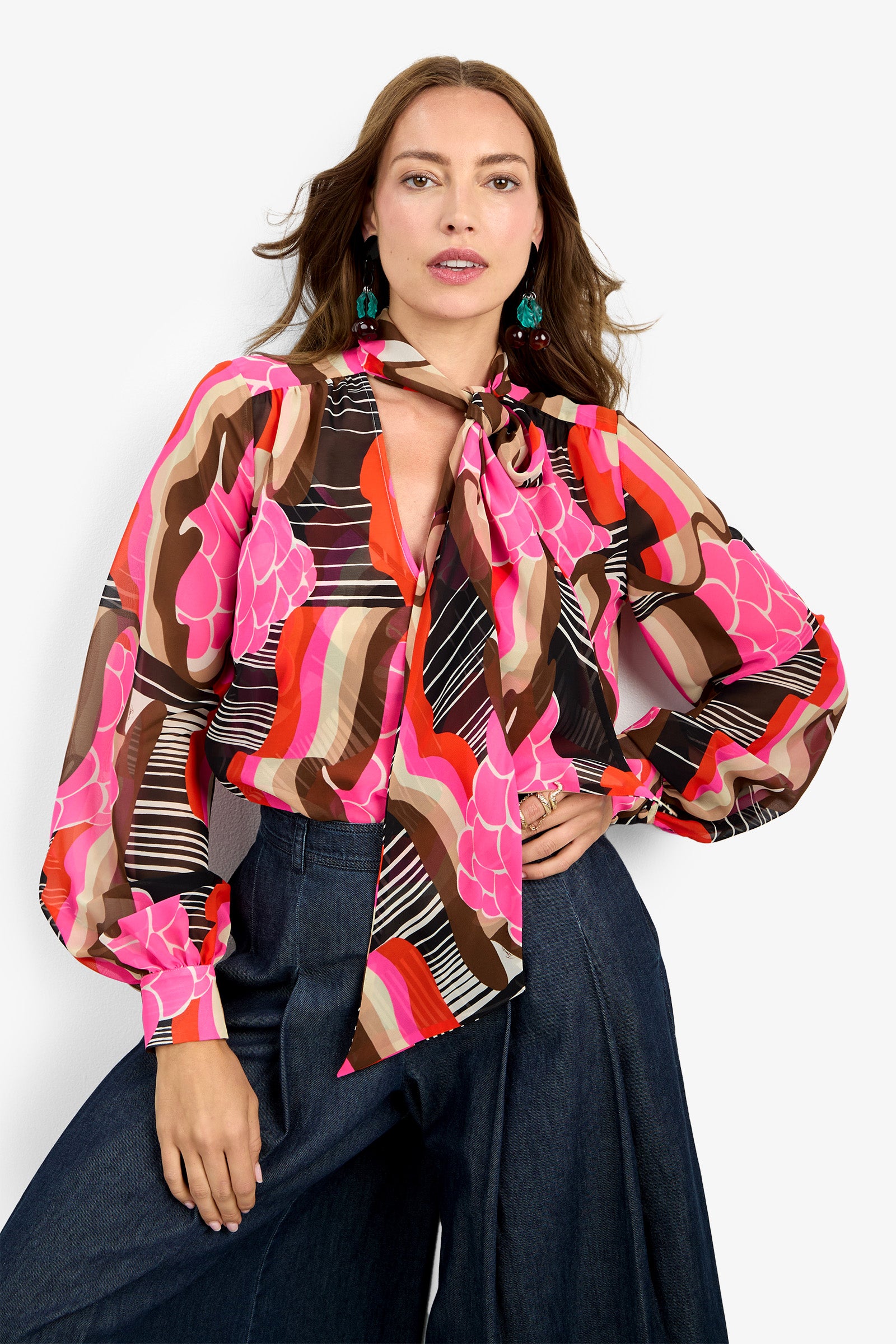 A woman wears the PIENZA TOP—a colorful, patterned blouse with pink, red, brown, and black designs and a necktie bow—paired with high-waisted wide-leg dark jeans. Her hair is loose and she accessorizes with green earrings.