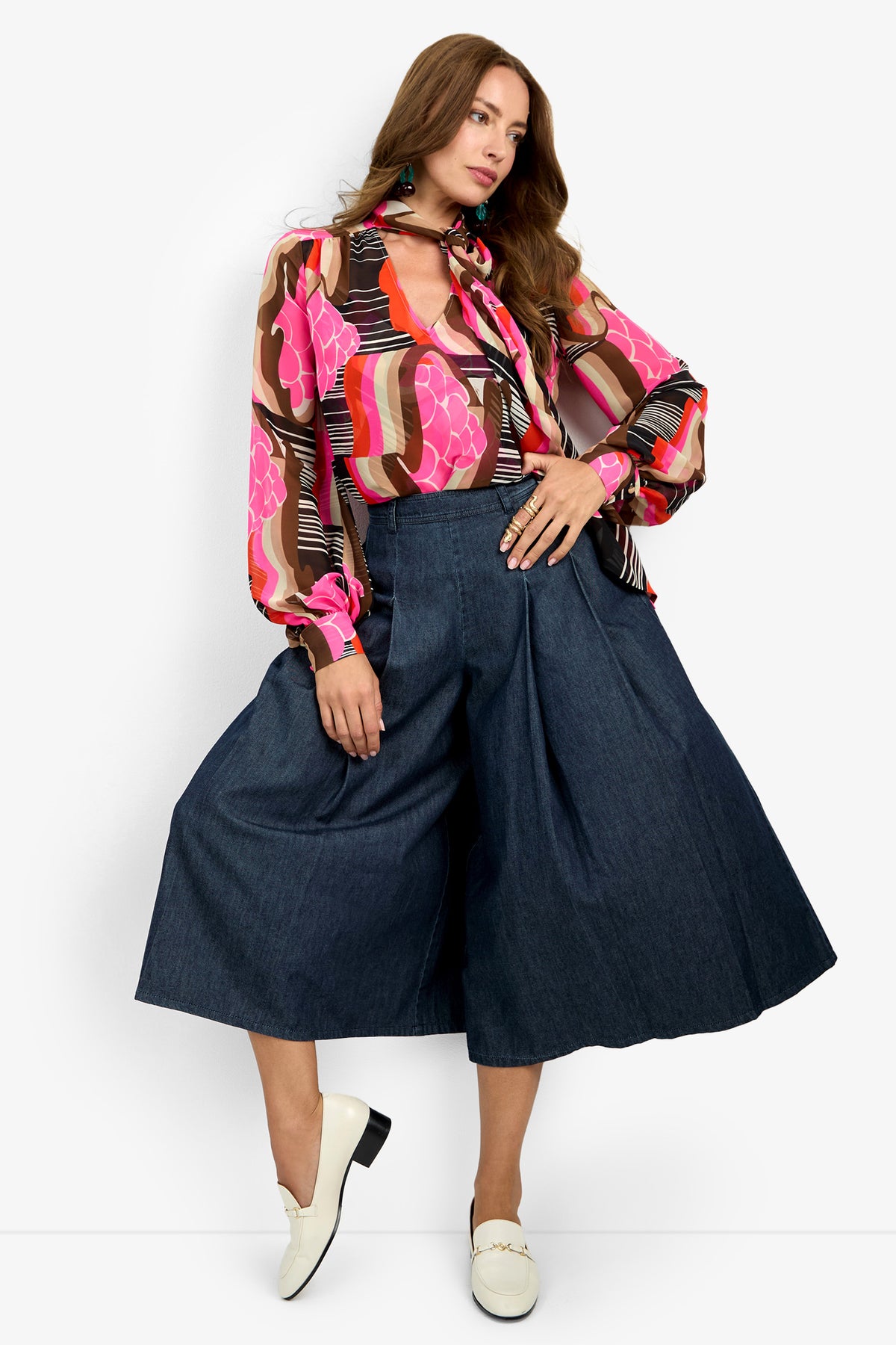 A woman poses in the PIENZA TOP, a colorful patterned blouse in pink, brown, and red tones, styled with high-waisted wide-leg denim culottes and white loafers.