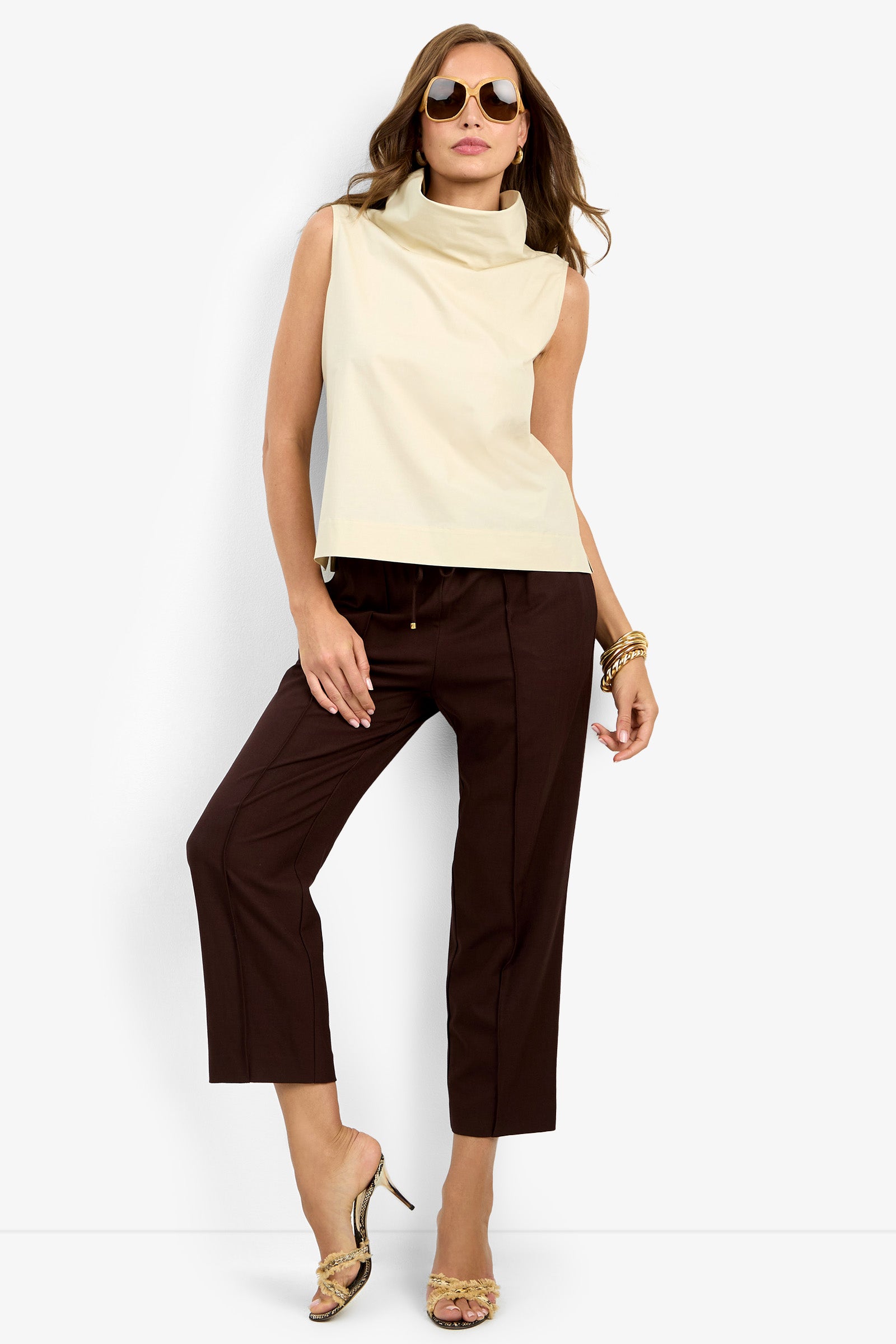 A woman in sunglasses wears the sleeveless beige CASTIGLIONE TOP, dark brown cropped pants, gold high-heeled sandals, and gold bracelets as she poses against a plain white background.