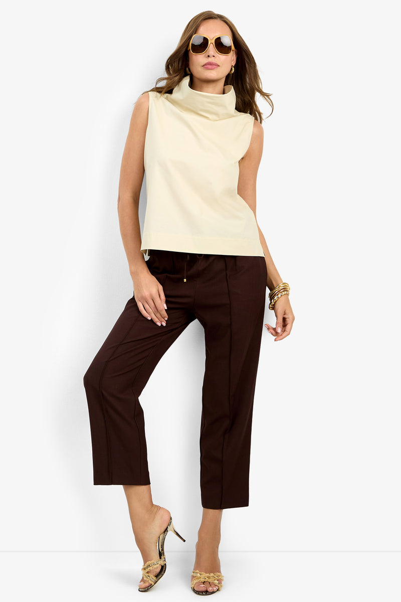 A woman in sunglasses wears the sleeveless beige CASTIGLIONE TOP, dark brown cropped pants, gold high-heeled sandals, and gold bracelets as she poses against a plain white background.