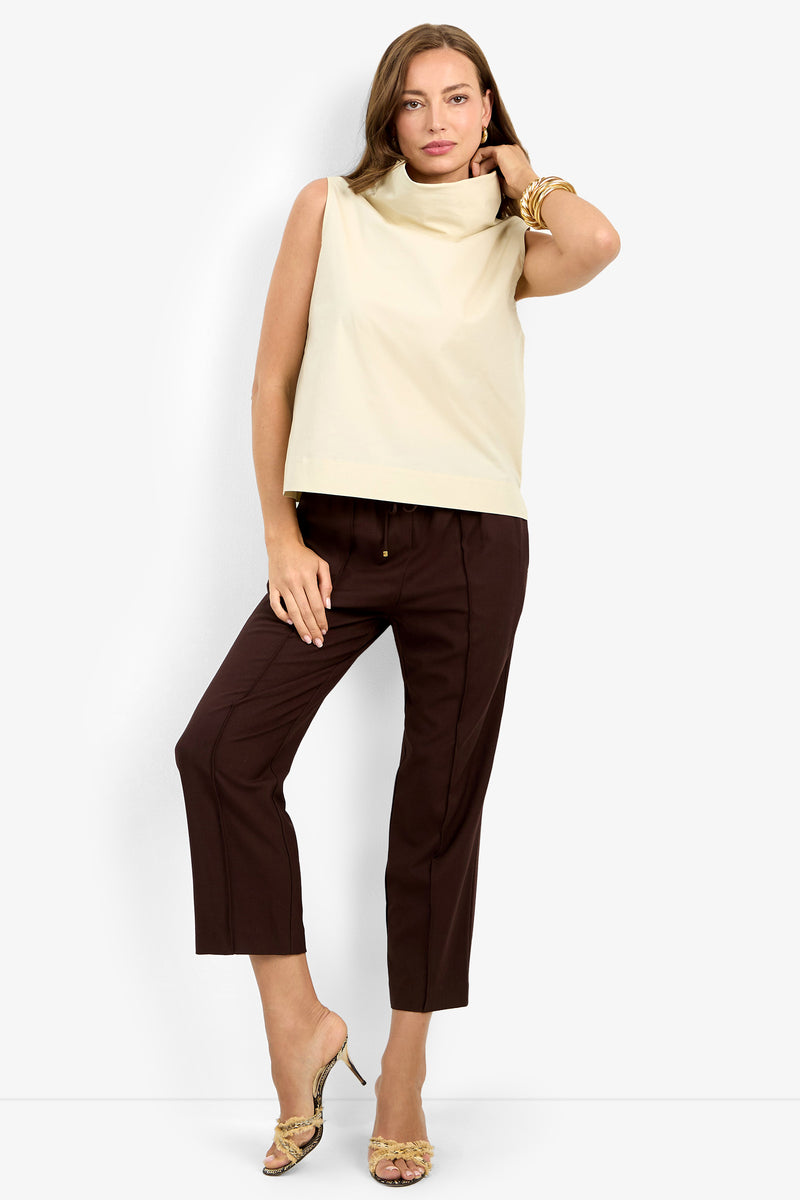 A woman poses in the CASTIGLIONE TOP, a sleeveless beige top with a wide collar, paired with brown cropped trousers, gold bracelets, and patterned high-heeled sandals against a plain white background.