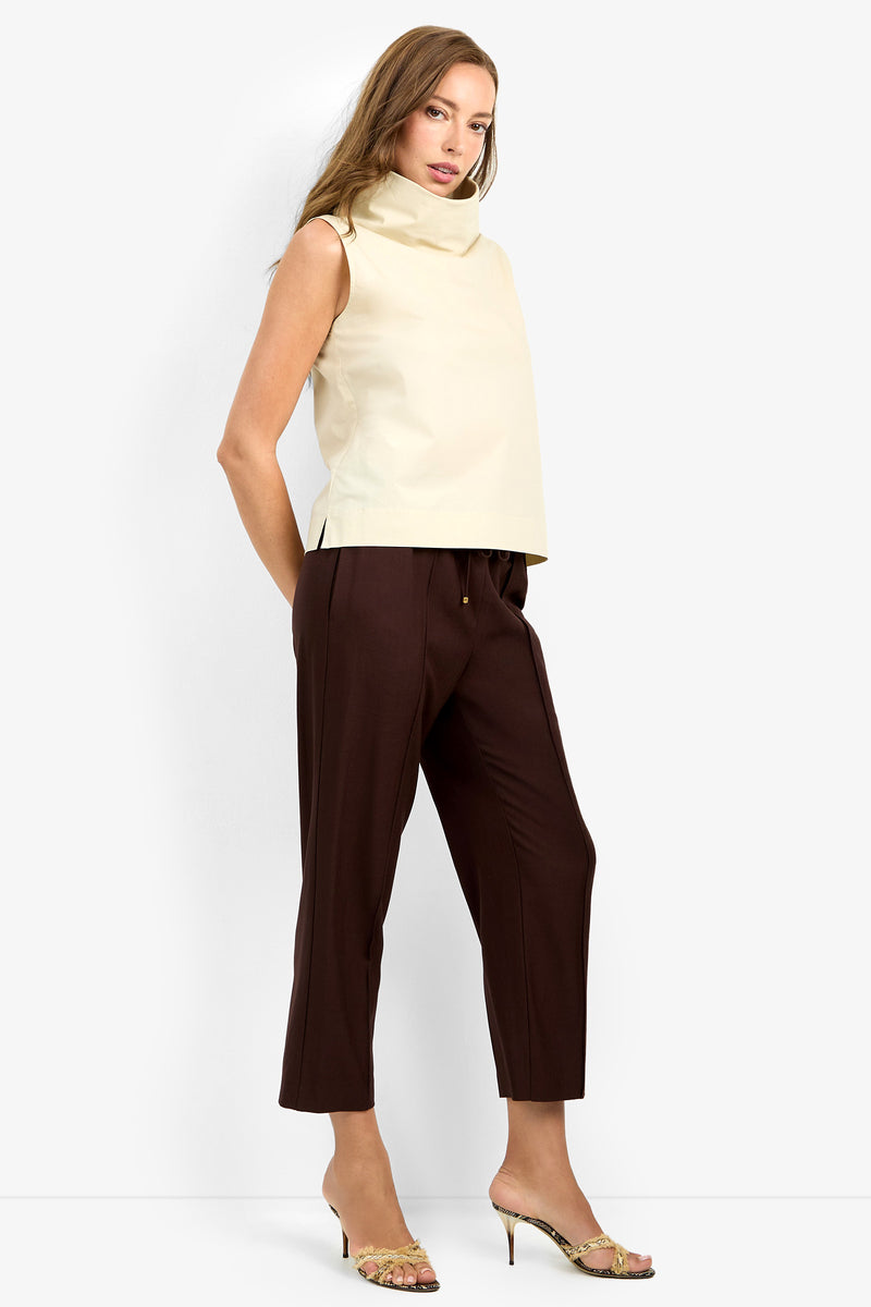 A woman with long hair poses confidently against a white background, wearing the sleeveless, high-neck CASTIGLIONE TOP in beige, paired with dark brown cropped trousers and gold-detailed open-toe heeled sandals.