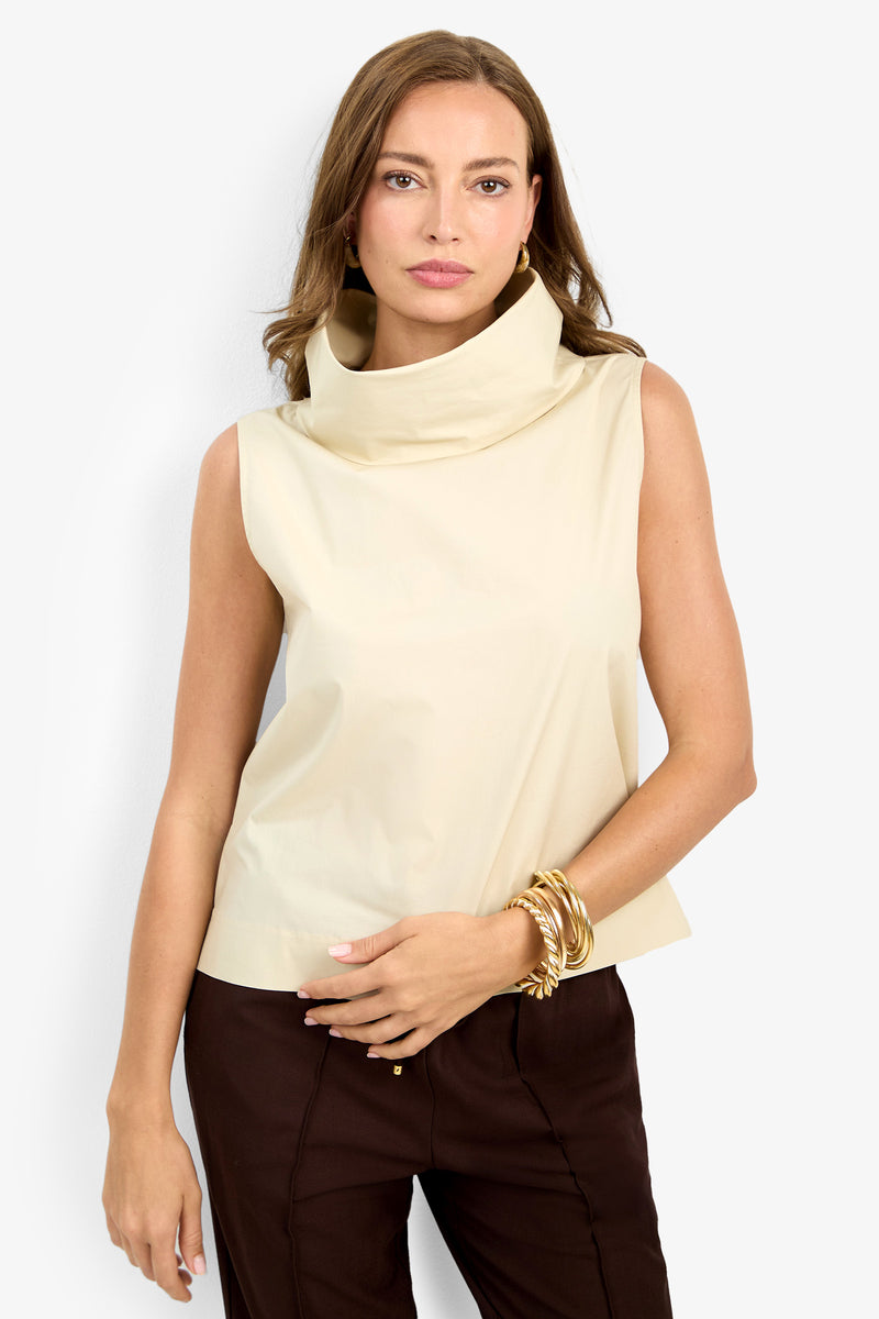 A woman with light brown hair models the CASTIGLIONE TOP, a sleeveless beige cowl-neck style, paired with dark pants and gold bangles, standing against a plain light background.