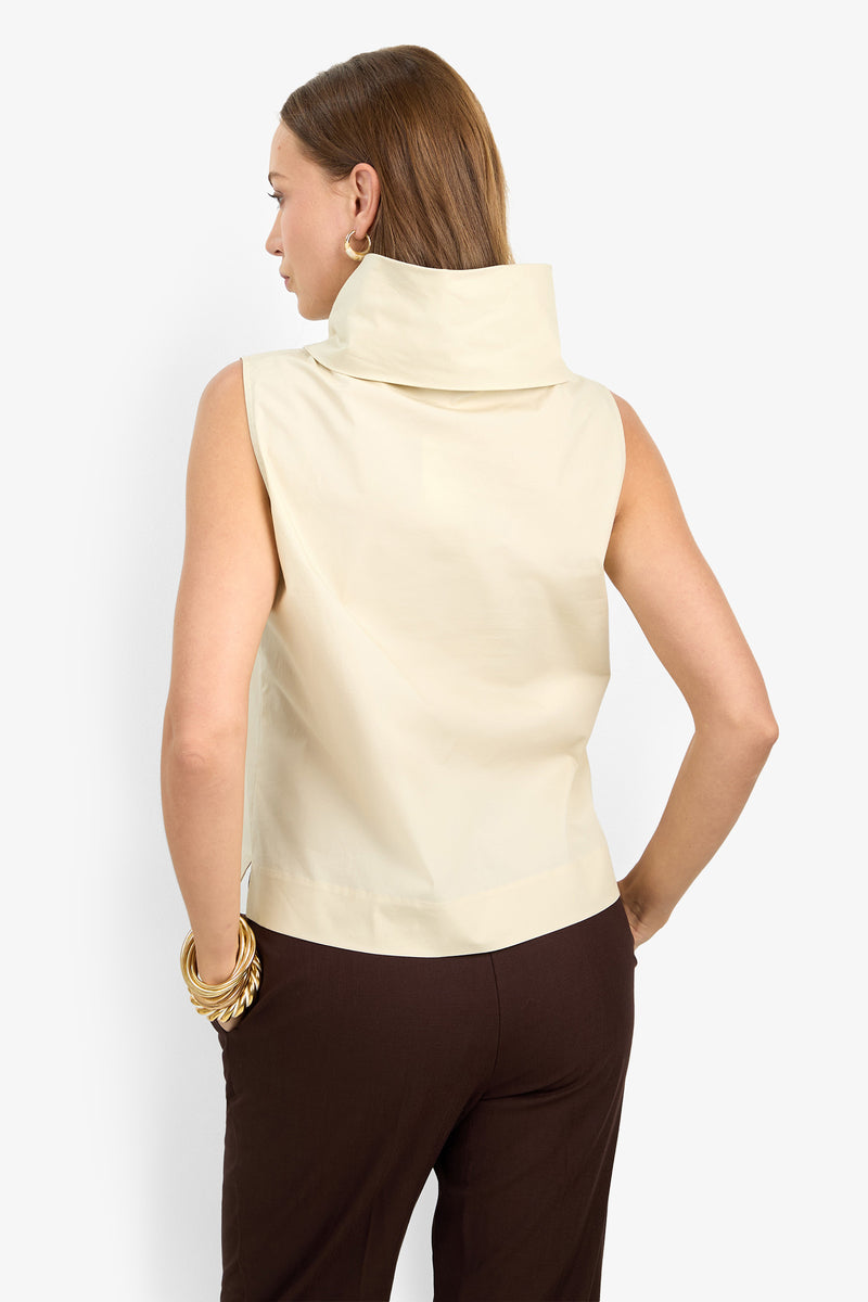 A woman with brown hair wears the CASTIGLIONE TOP, a sleeveless high-collared cream blouse, paired with dark brown pants. She faces away from the camera, sporting large gold hoop earrings and chunky gold bracelets.