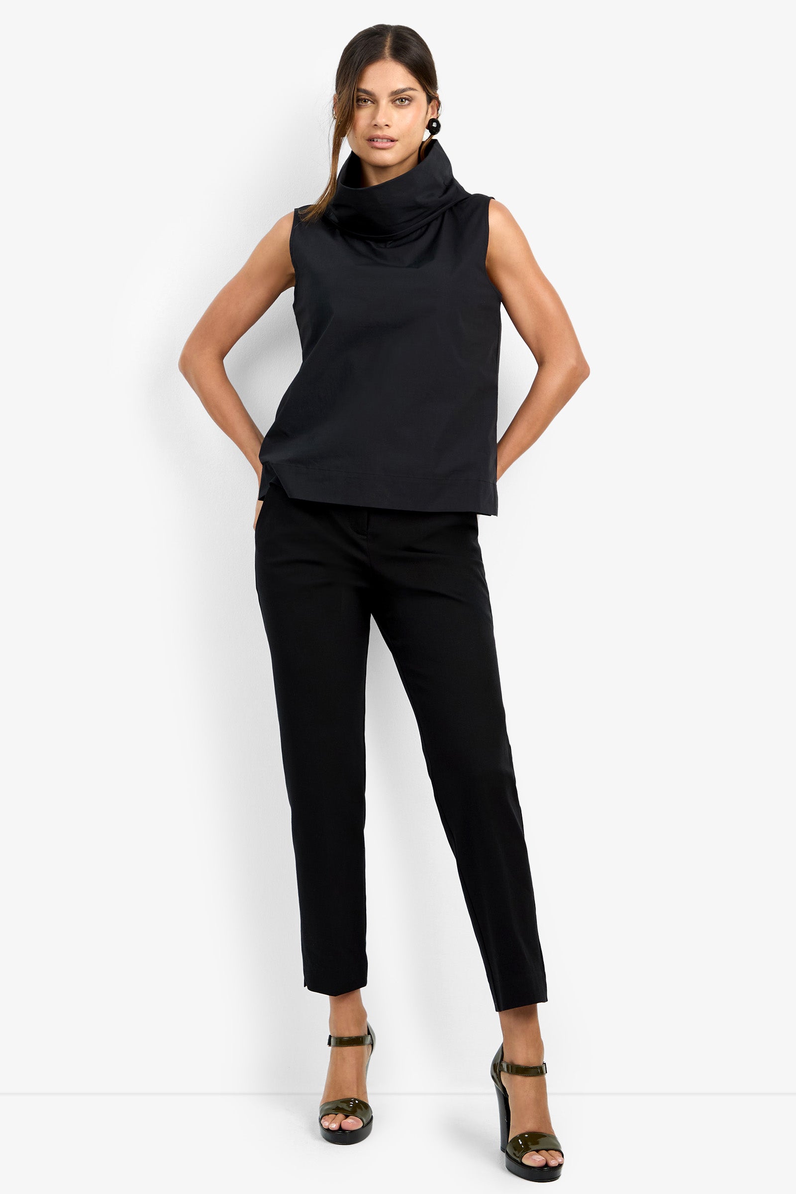 A woman stands confidently against a white background, wearing the CASTIGLIONE TOP—a sleeveless black cowl-neck piece—paired with black tailored pants, black heeled sandals, large round earrings, and her hair in a loose ponytail.