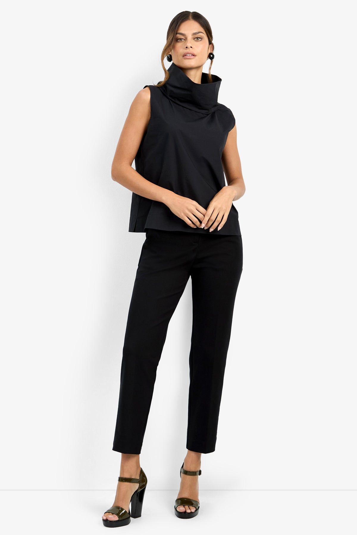 A woman poses against a white background wearing the CASTIGLIONE TOP—a sleeveless black high-collar top—paired with black pants, dark heeled sandals, and large round earrings. One hand rests on her thigh, the other at her side.