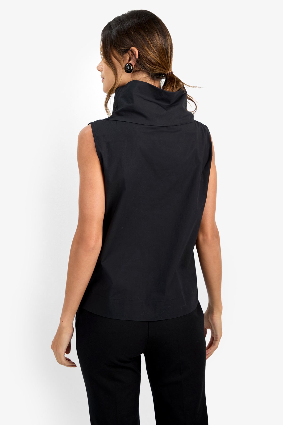 A woman faces away, wearing the CASTIGLIONE TOP—a sleeveless black top with a wide, high collar—paired with black pants and circular earrings. Her brown hair is styled back against a plain white background.