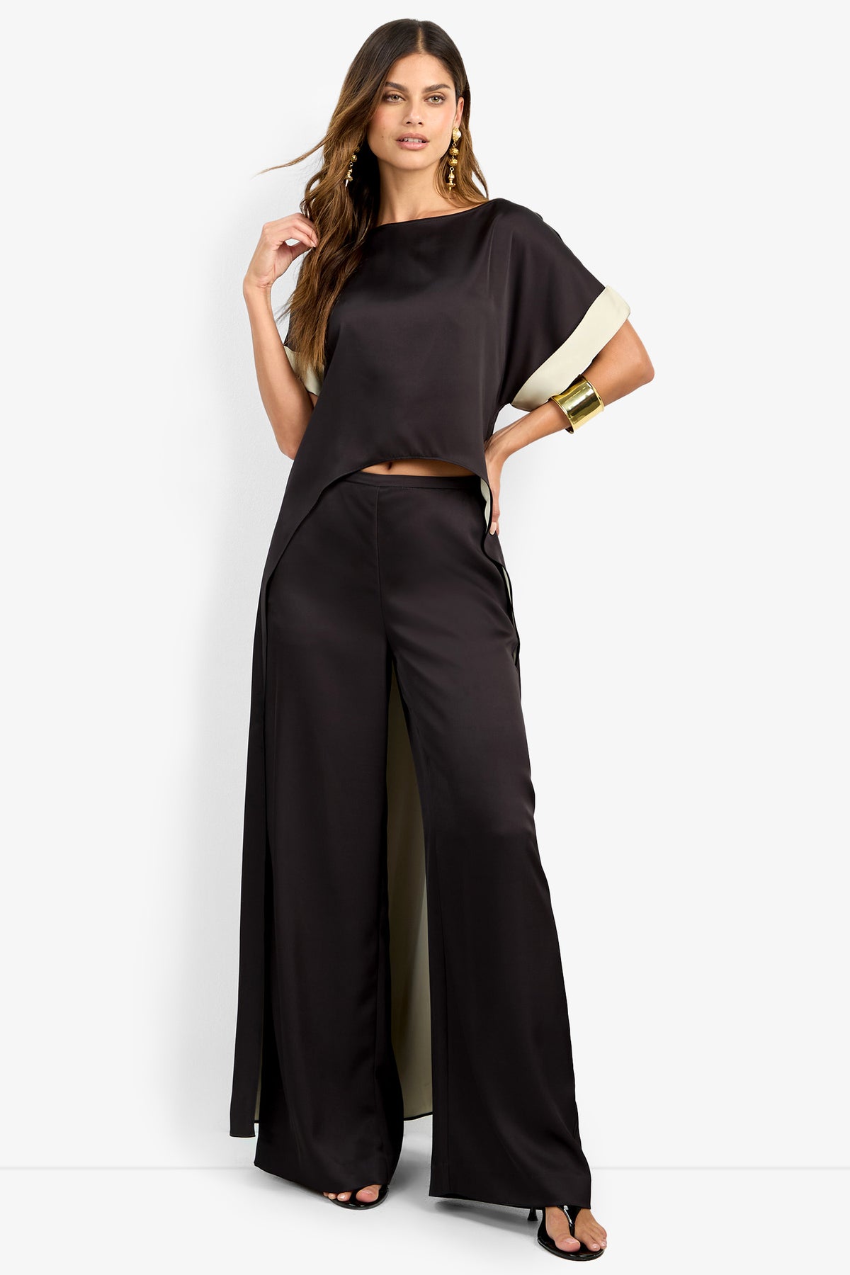 A woman poses in the MATTIAH TOP, a loose black satin blouse with wide sleeves, paired with matching wide-leg pants. She completes the look with gold earrings, a cuff bracelet, and black heels against a white background.