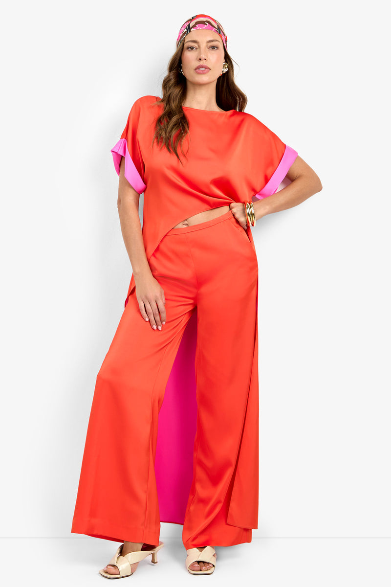 A woman stands confidently against a white background wearing the bright orange MATTIAH TOP and wide-leg pants with pink accents, beige heeled sandals, a chunky bracelet, and a colorful headband.