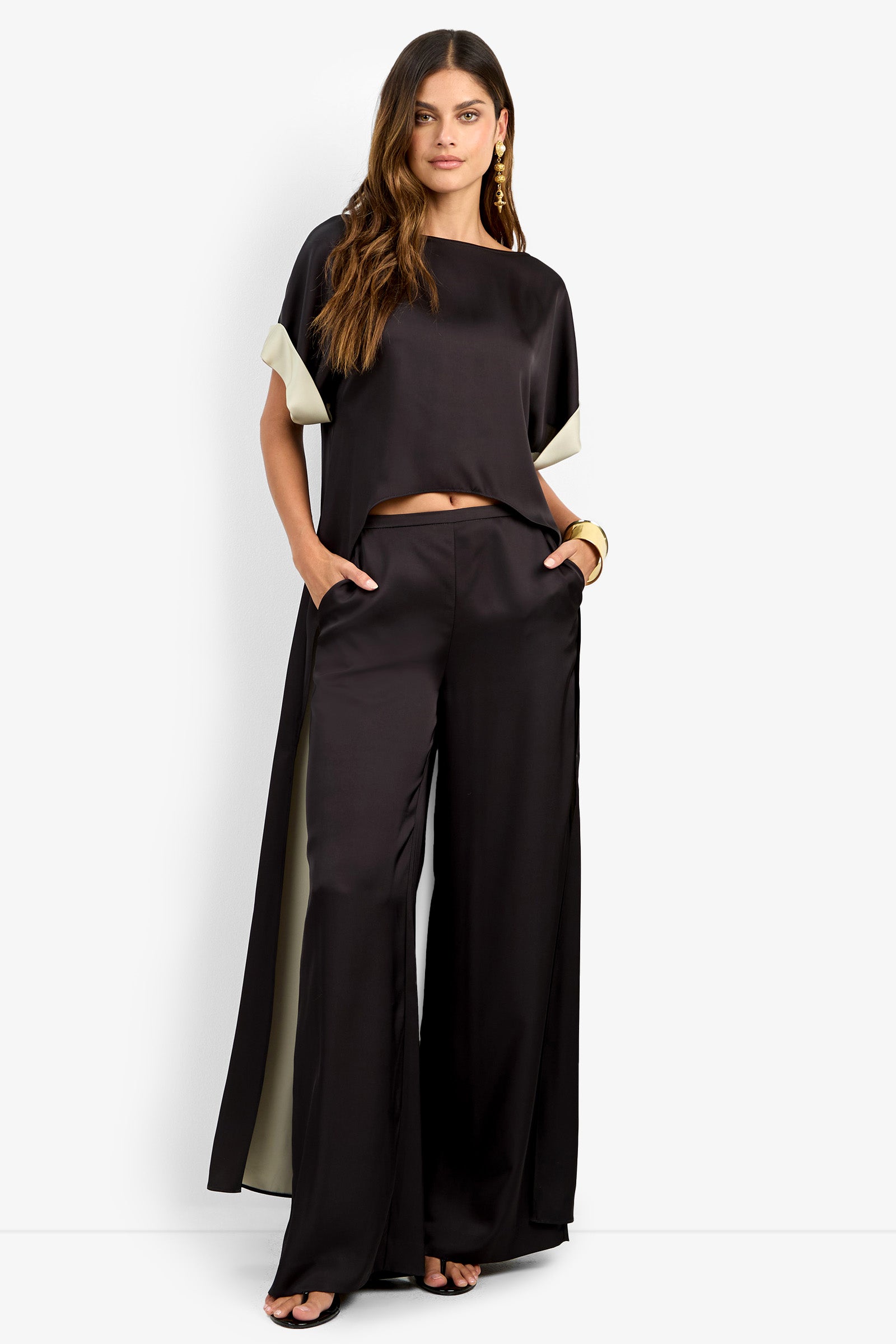 A woman wears the loose-fitting black MATTIAH TOP with wide-leg satin pants, short sleeves, and light sleeve lining. She poses with hands in pockets and black heels.