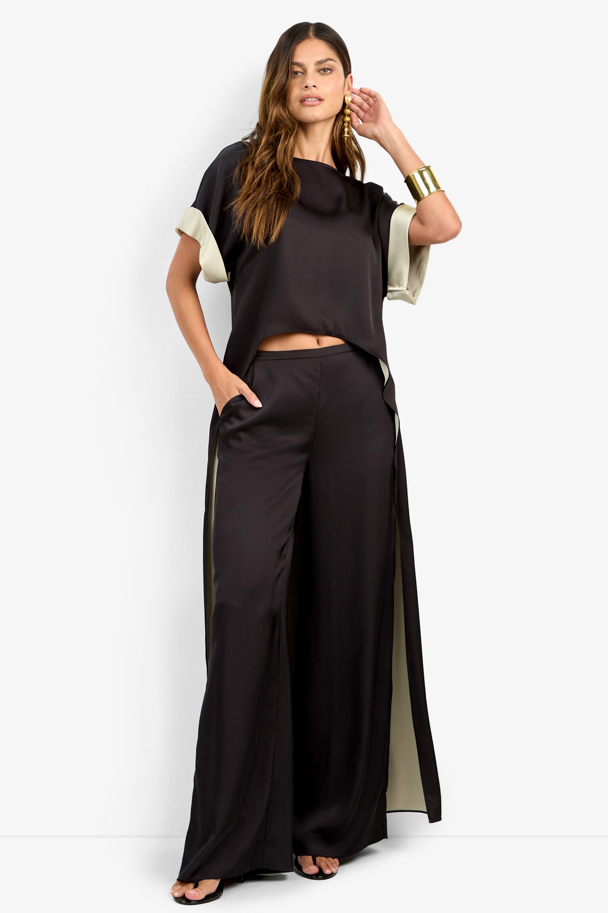 A woman wears the MATTIAH TOP, a loose black satin short-sleeve blouse, paired with matching wide-leg pants featuring cream side panels. She accessorizes with a gold cuff bracelet and black sandals, her long wavy hair cascading down.