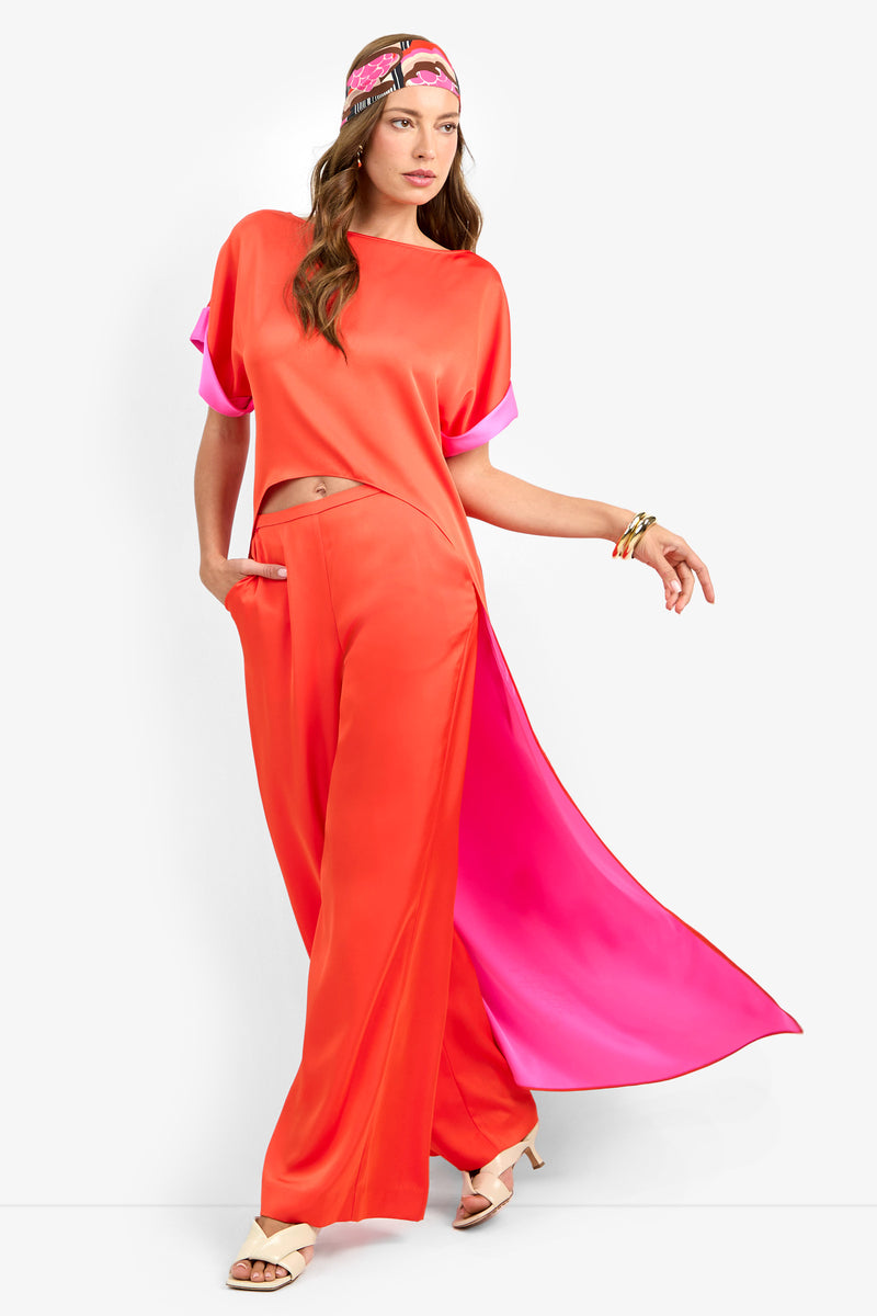A woman confidently poses in the MATTIAH TOP—an orange top with pink accents—paired with matching wide-leg pants, a coordinating pink-orange headscarf, bangles, and beige heeled sandals.