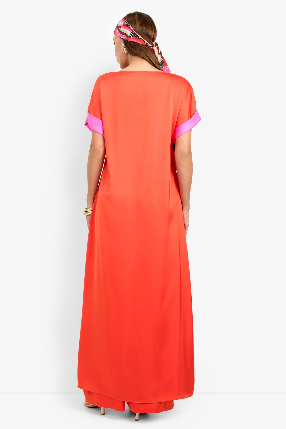 A person stands facing away in the MATTIAH TOP—a long, loose, bright orange piece with pink cuffs—paired with wide pants and a colorful headscarf. The stylish, vibrant look is set against a plain white background.