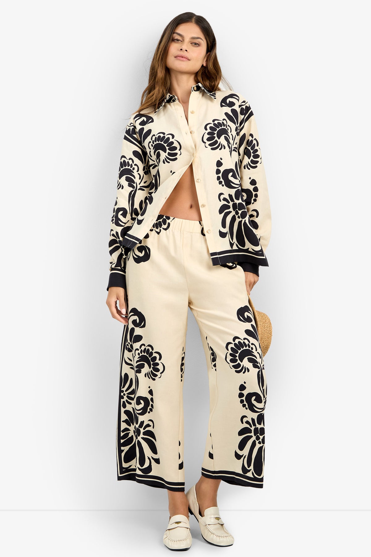 A woman wears the NIVARA TOP, a cream shirt with bold black floral patterns, paired with matching wide-leg pants. She stands against a white background, holding a straw hat and wearing white loafers.