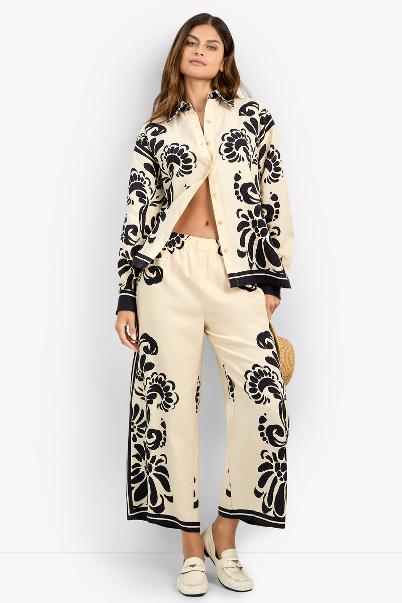 A woman wears the NIVARA TOP, a cream shirt with bold black floral patterns, paired with matching wide-leg pants. She stands against a white background, holding a straw hat and wearing white loafers.