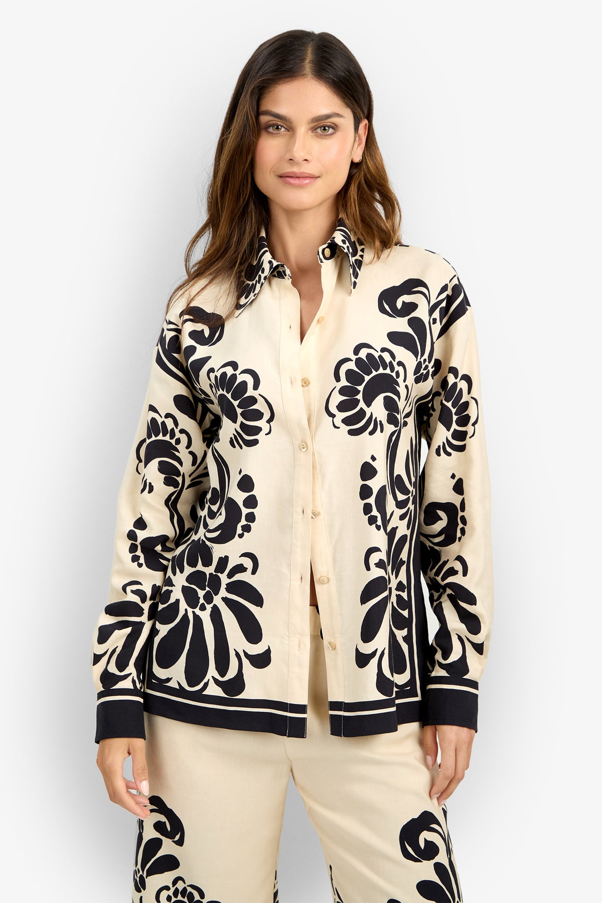 A woman with long hair and a neutral expression poses against a plain light background wearing the NIVARA TOP, a beige button-up shirt with bold black floral patterns, styled with matching pants.