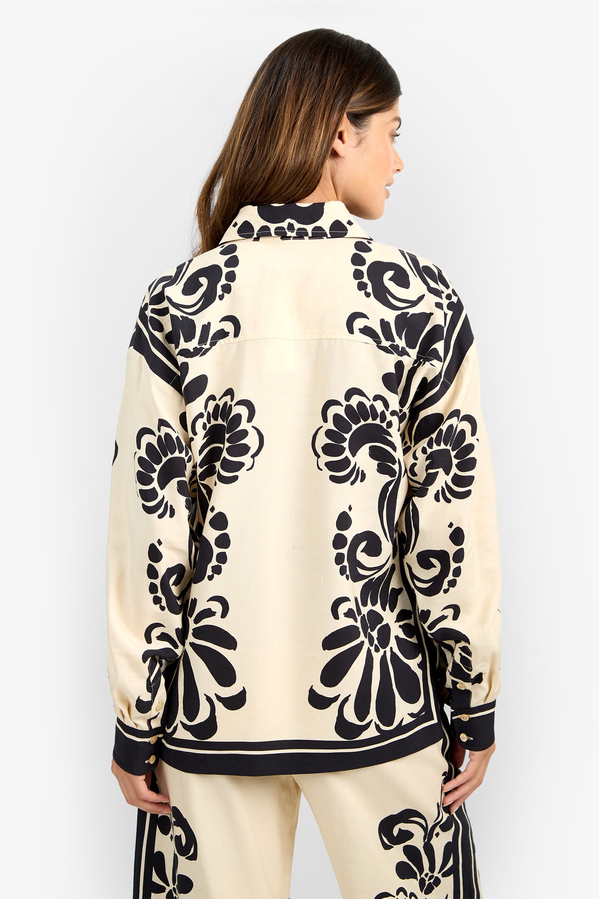 A woman with long brown hair is seen from the back wearing the NIVARA TOP, a cream-colored shirt featuring bold black floral patterns, paired with matching pants against a plain white background.