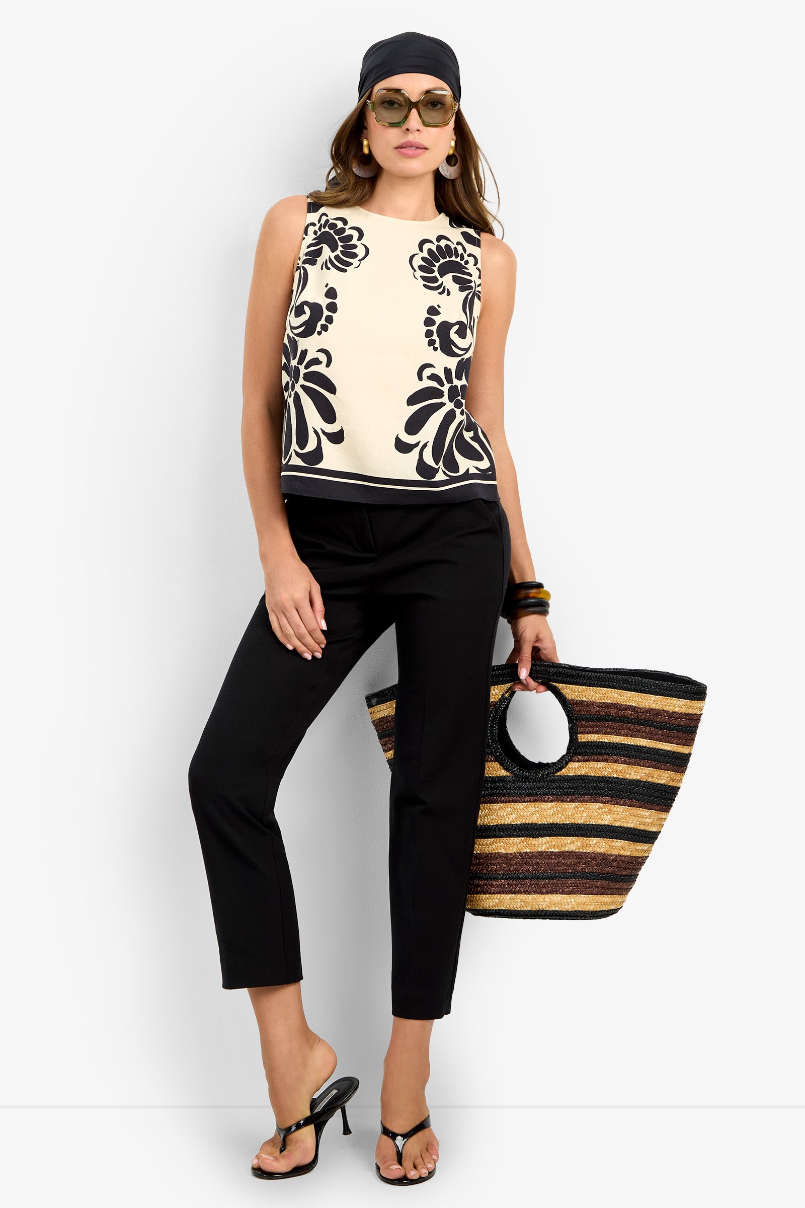 A woman stands confidently against a plain background, wearing the sleeveless patterned MALEMA TOP, paired with black pants, sandals, headscarf, sunglasses, and carrying a large striped woven bag.