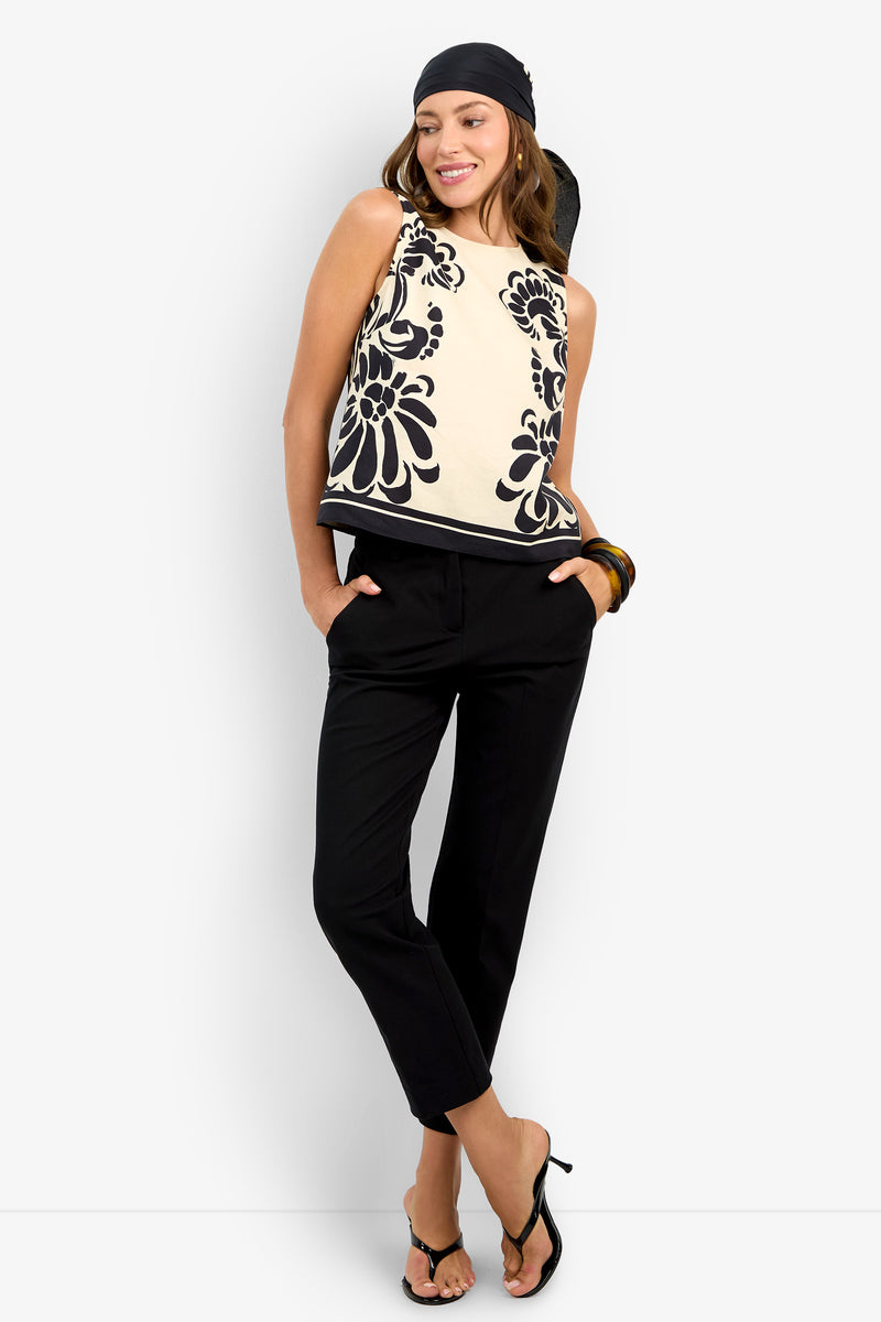A woman stands against a white background, smiling with her hands in her pockets, wearing the MALEMA TOP—a sleeveless cream top with black floral patterns—paired with a black headscarf, black pants, heeled sandals, and a chunky bracelet.